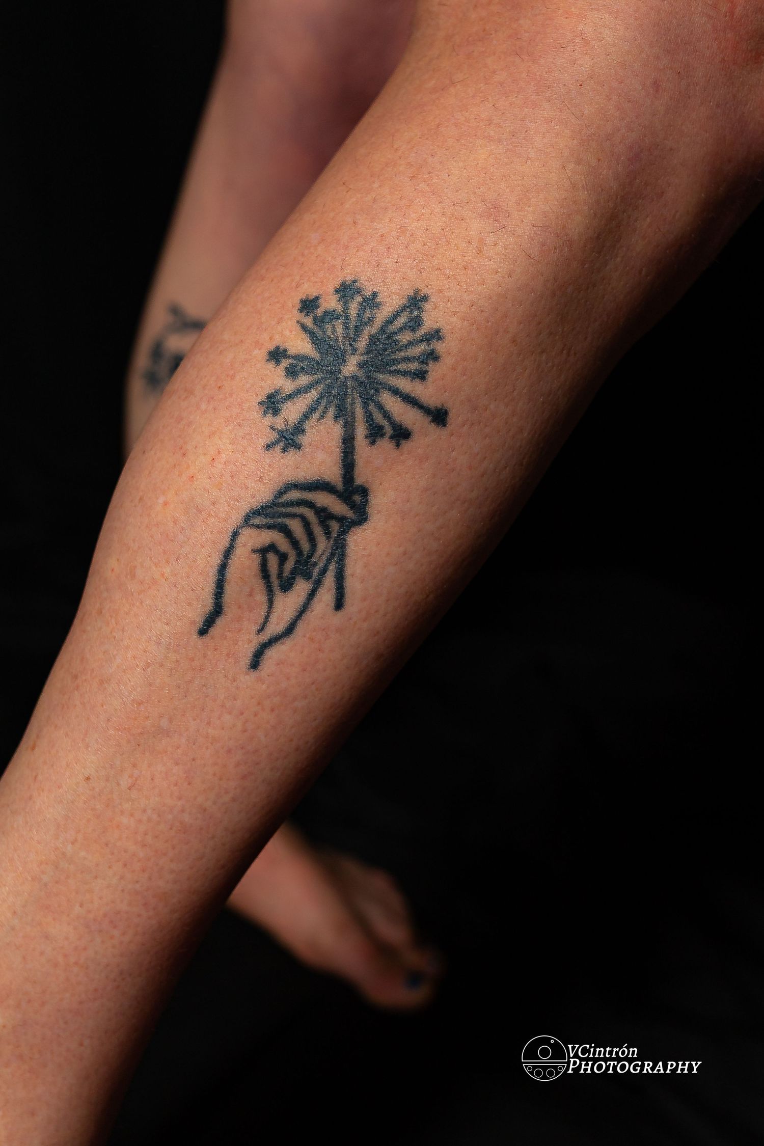 Black tattoo of a hand holding a delicate dandelion flower design on someone's forearm.