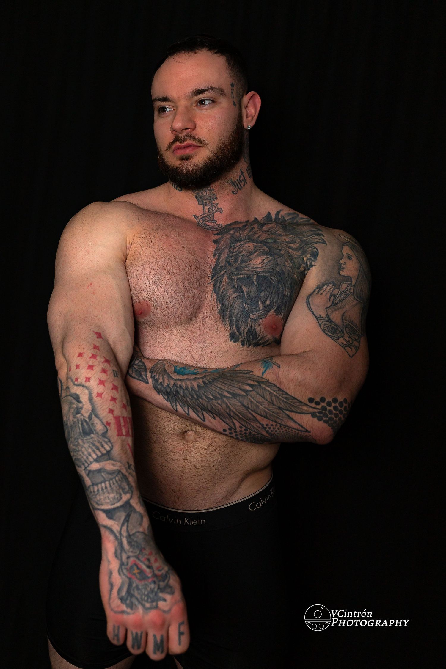 Shirtless muscular man with numerous artistic tattoos including chest and arm pieces poses against dark background.