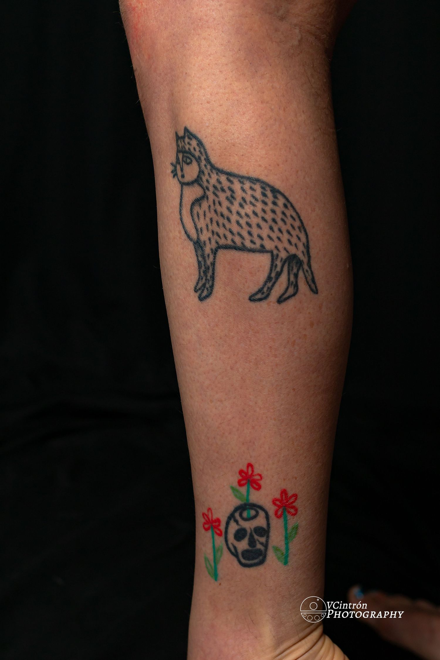 Minimalist black linework tattoo of a spotted wildcat above small red flowers and a circle design on someone's arm.