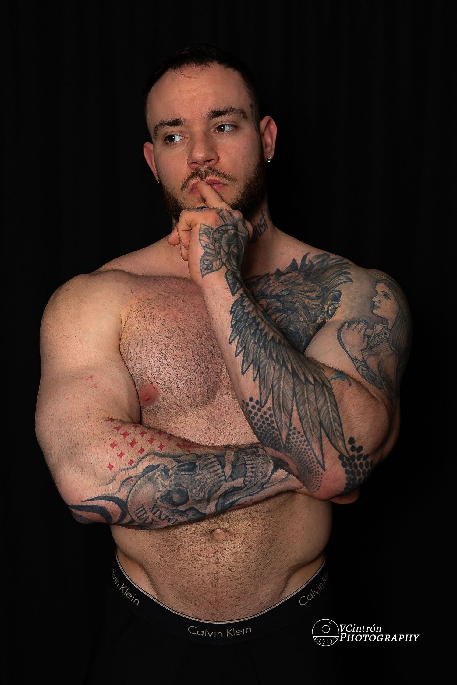 Shirtless muscular person with elaborate tattoo sleeves poses thoughtfully against dark background wearing Calvin Klein underwear.