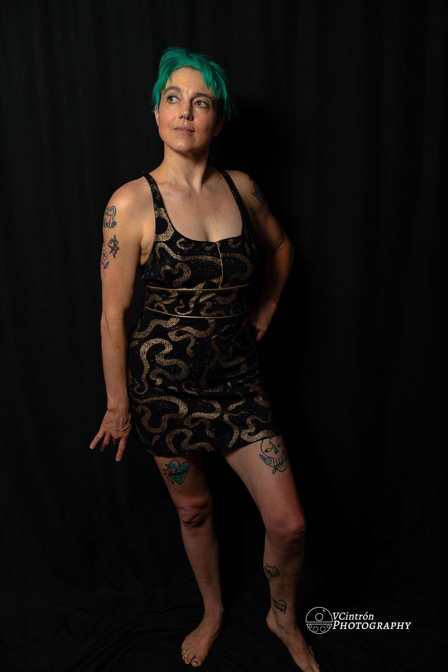 Studio portrait against black background showing figure with green hair in black patterned dress with visible tattoos.