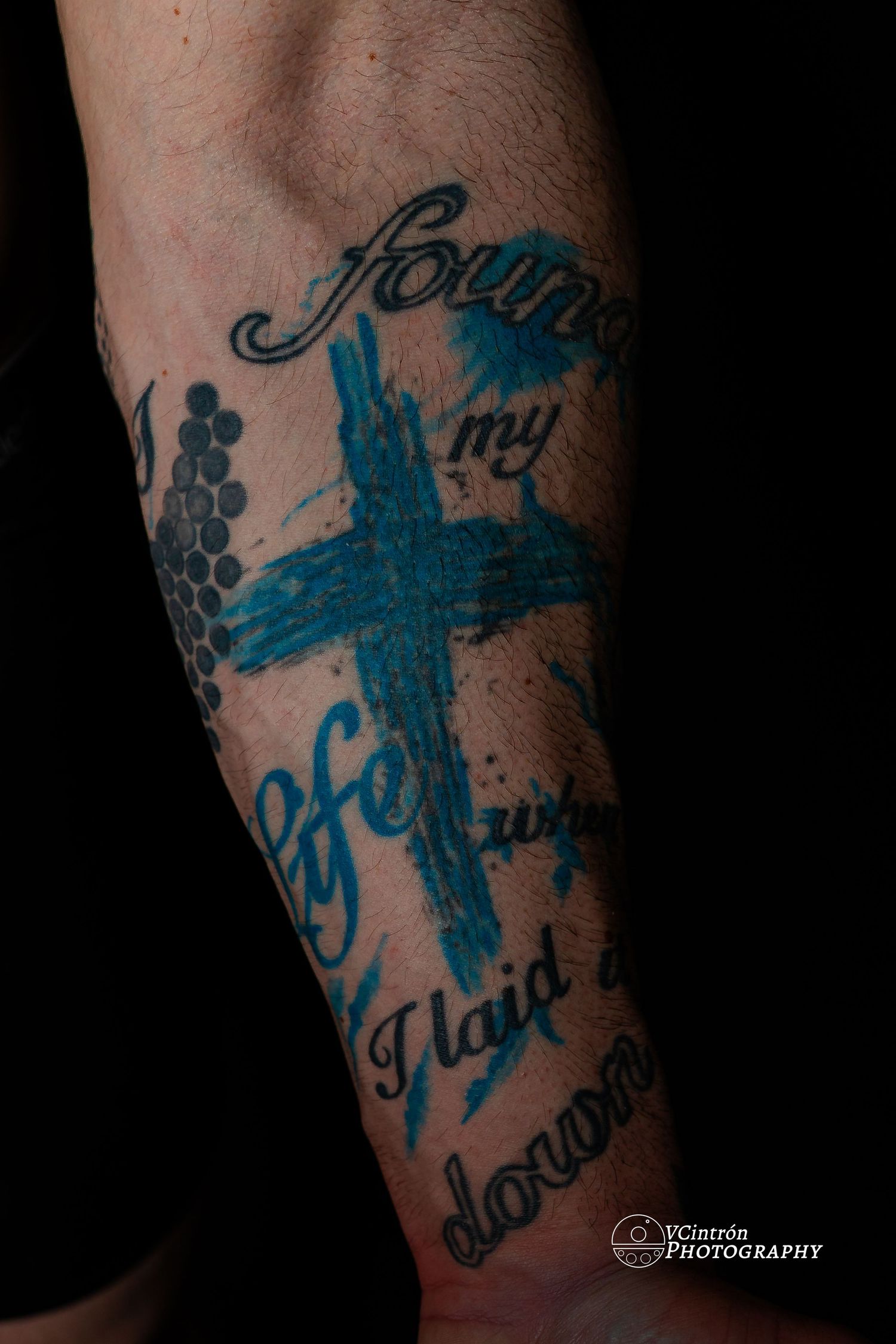 Close-up of a blue cross tattoo with script lettering and decorative elements on someone's arm.