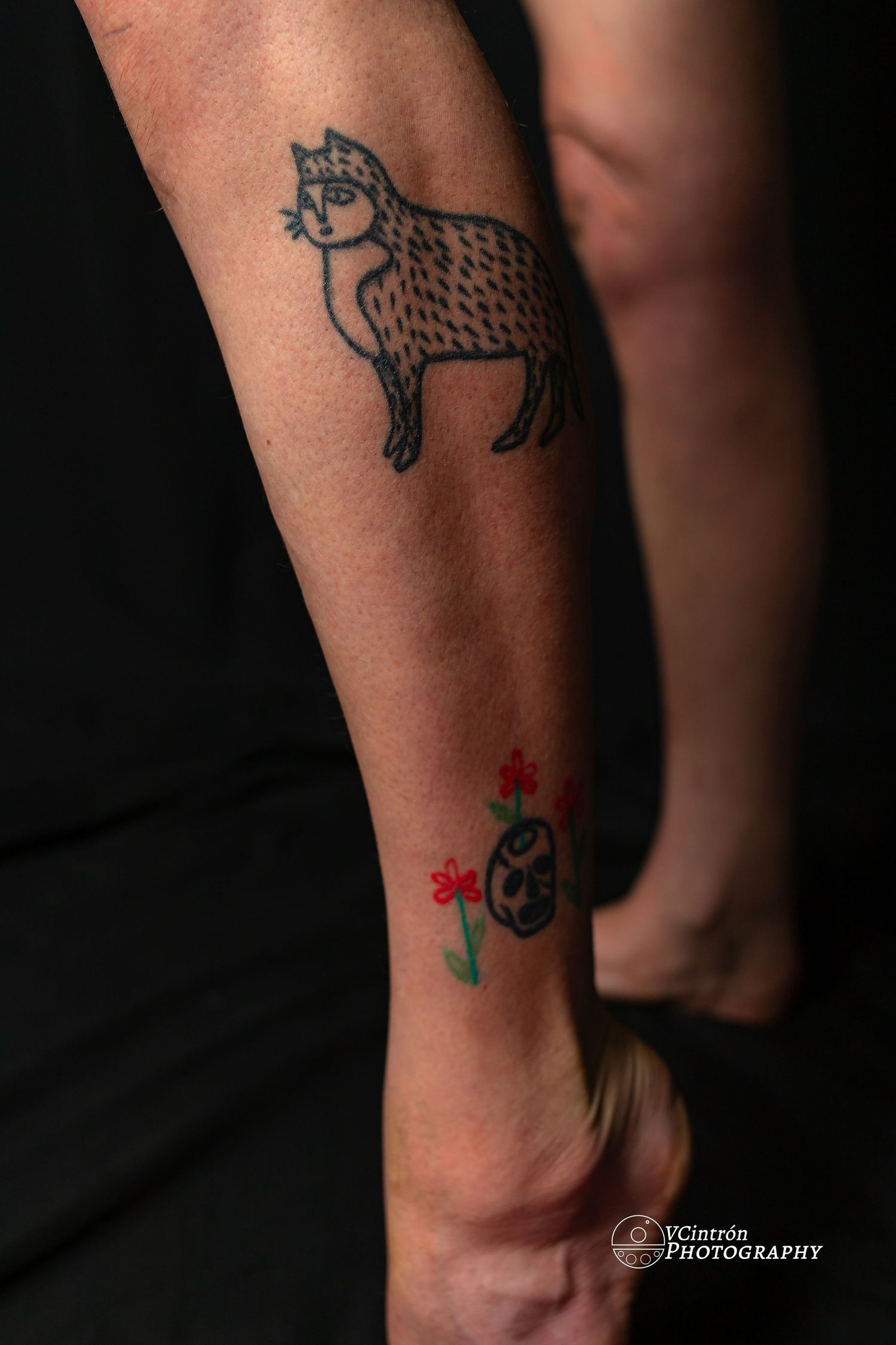 Two tattoos on an arm showing a decorative wolf design and small red flowers with a black circular symbol below.