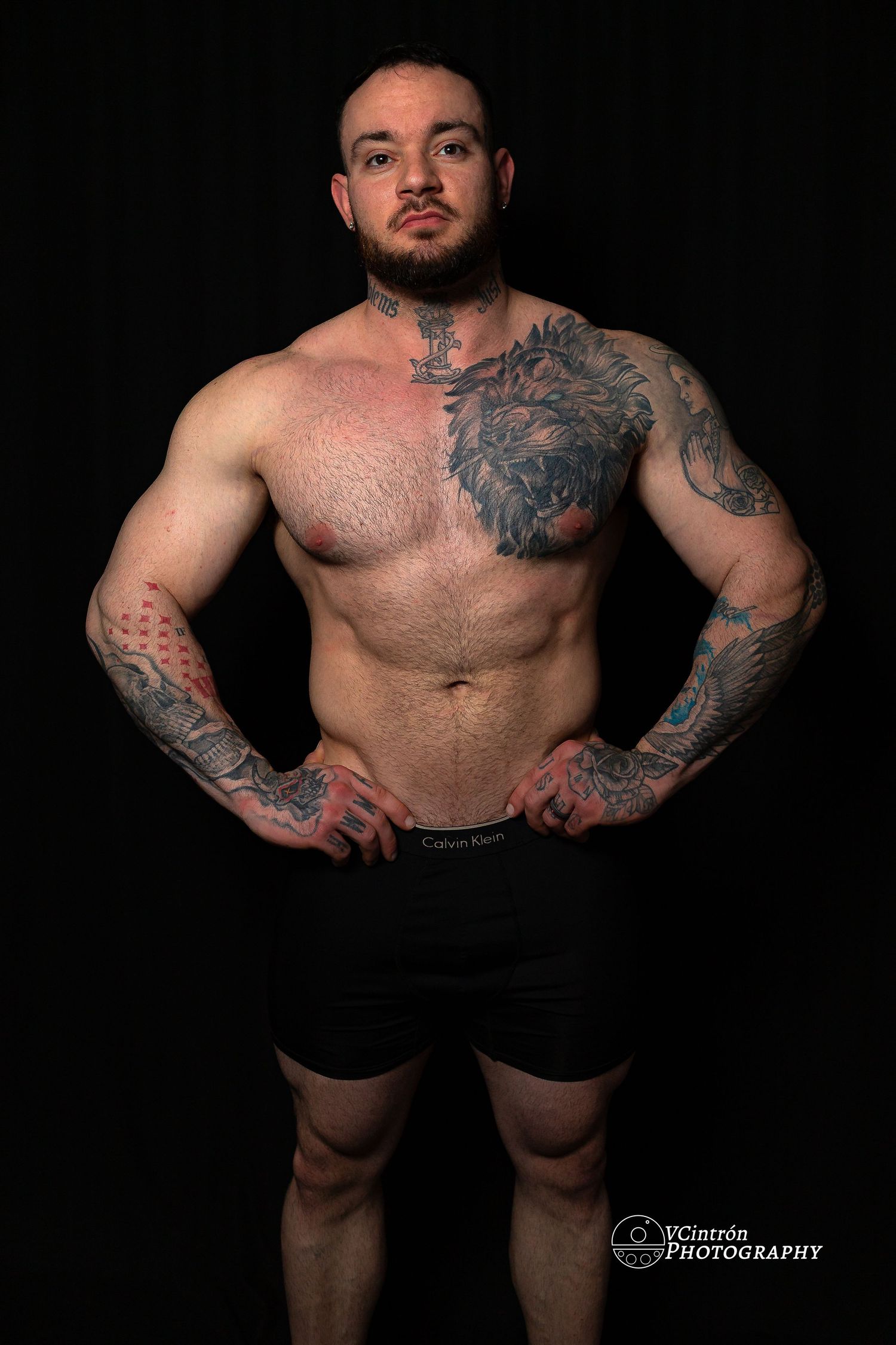 Muscular shirtless person with intricate tattoos on dark background wearing black shorts in dramatic studio lighting.
