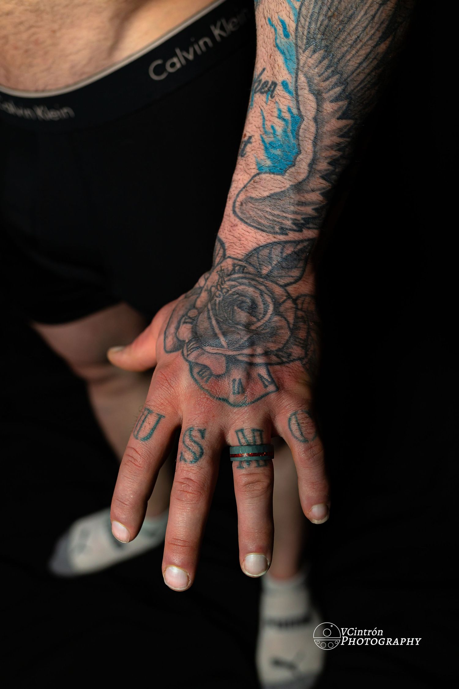 Hand with Calvin Klein clothing visible shows detailed rose and wing tattoo artwork in black and blue ink, with finger tattoos.