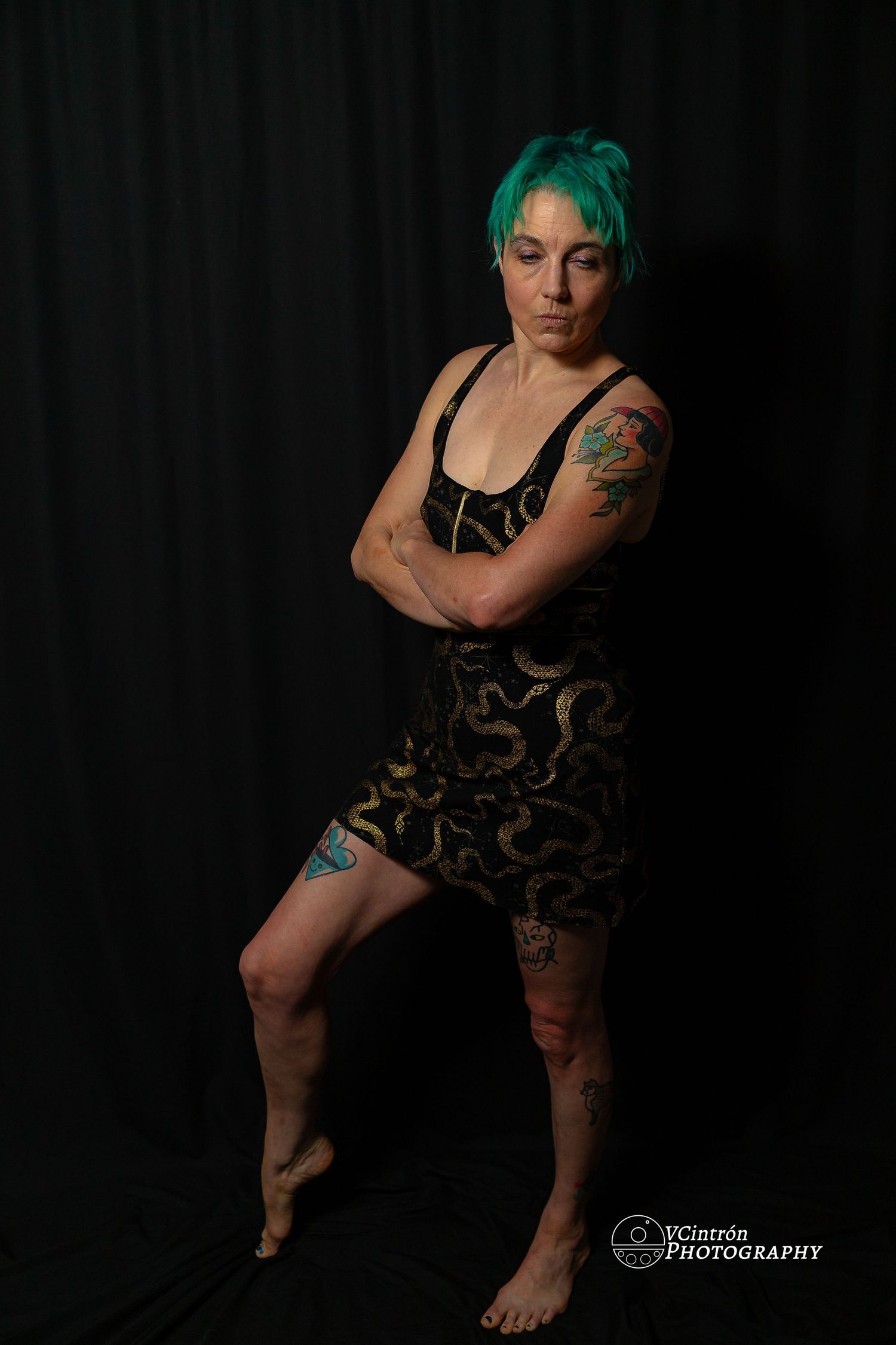 A figure with green hair poses against a black background wearing a patterned short dress and displaying visible tattoos.