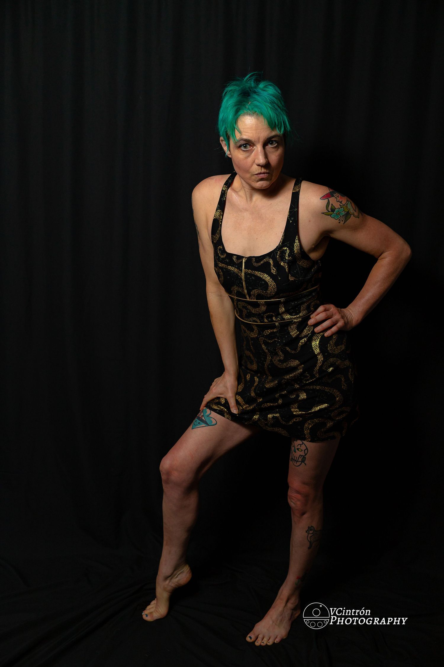 Studio portrait of someone with bright green hair wearing a black camo dress against a dark background.