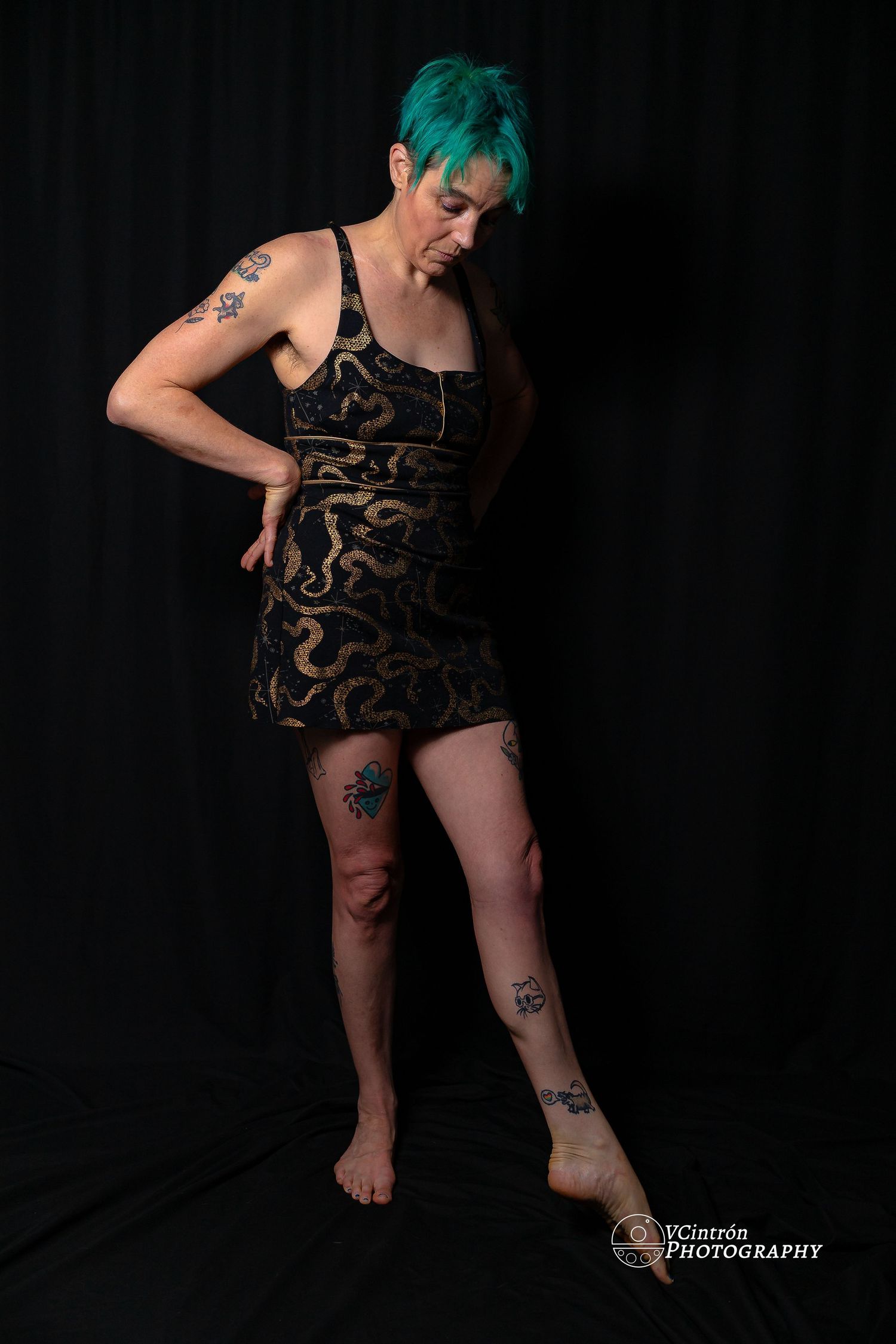 Figure wearing black and gold patterned dress with green hair poses dramatically against dark background showing tattoos.
