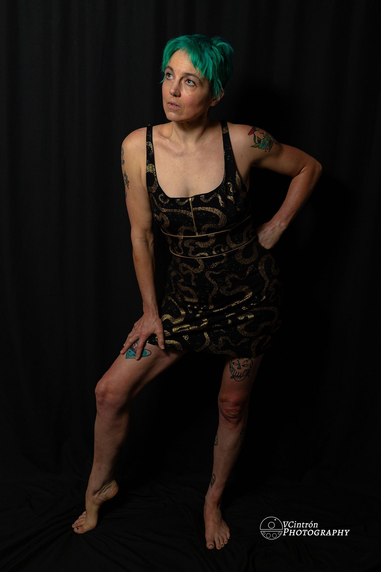 A figure in a leopard print dress and green hair poses dramatically against a black background in an artistic portrait.