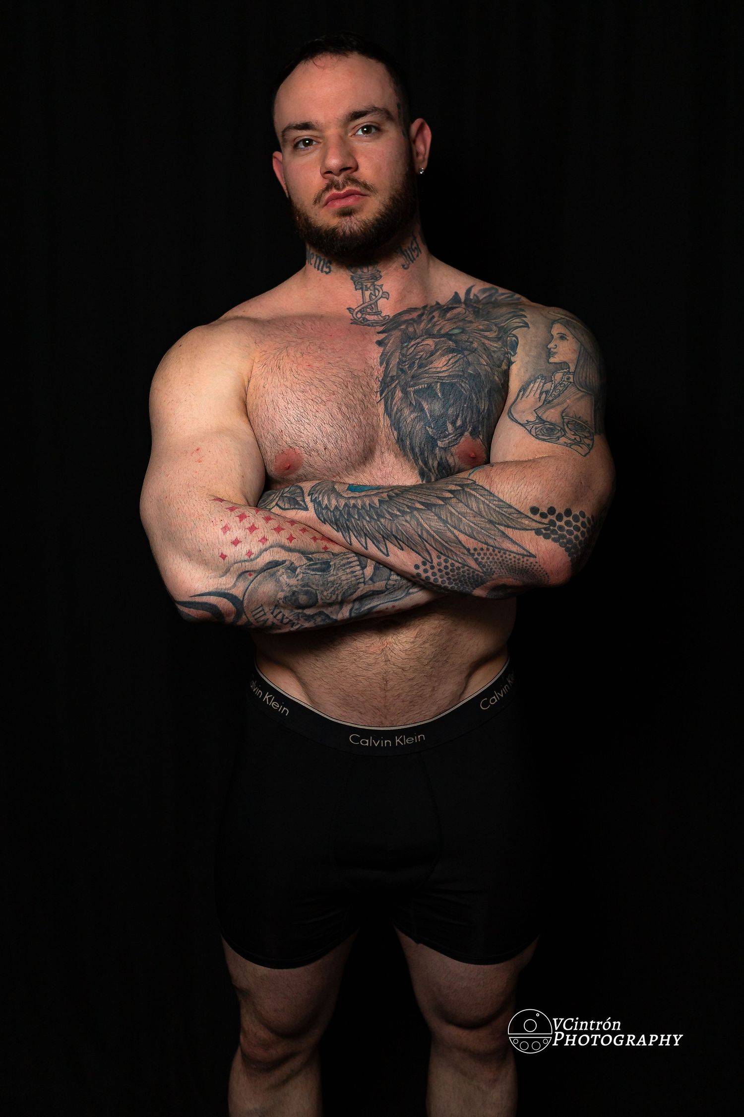 Studio portrait against black background showing muscular shirtless person with detailed sleeve tattoos in black shorts.