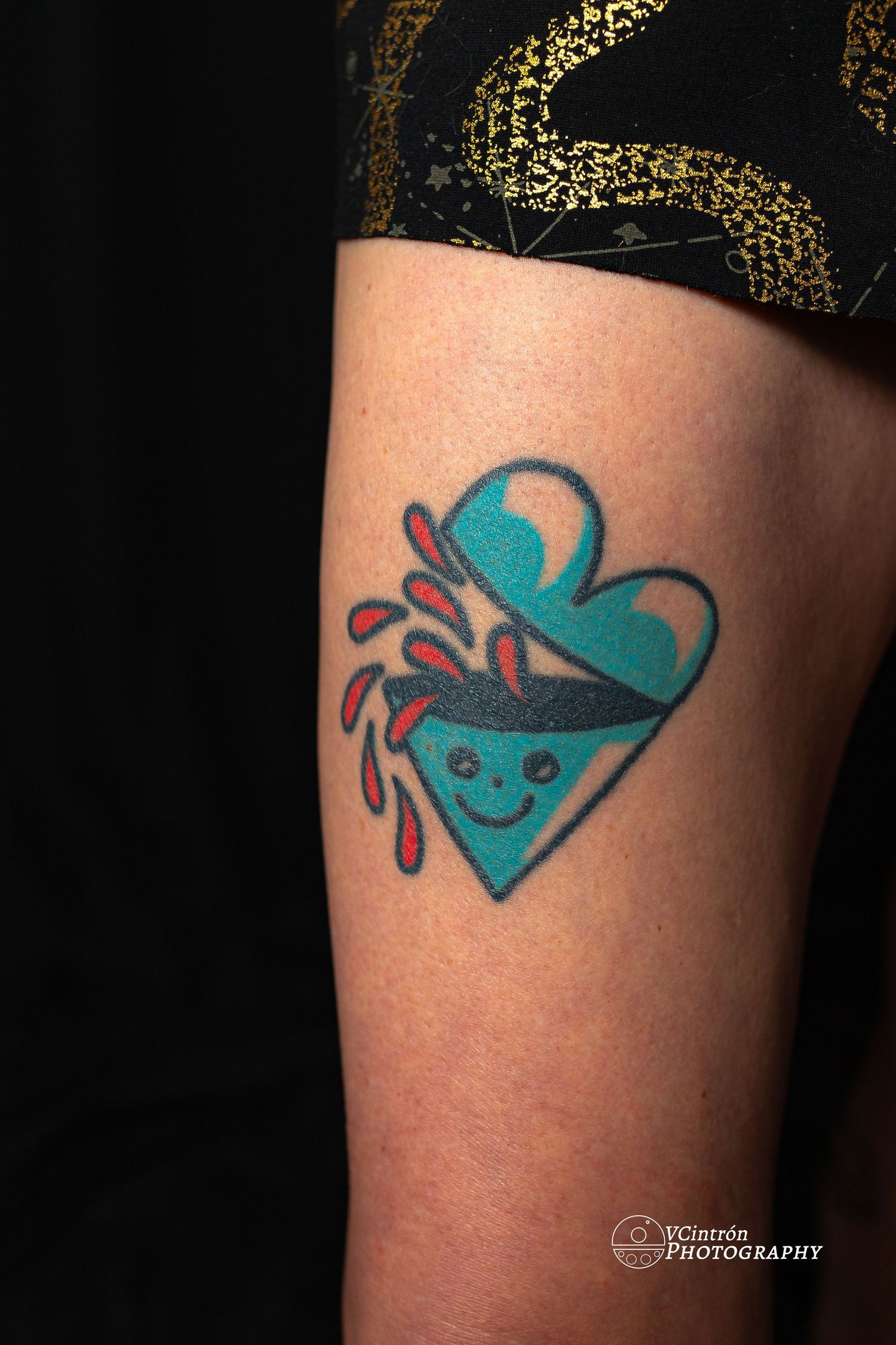 Small teal and red geometric heart tattoo with a smiling face and dripping liquid design on an arm.
