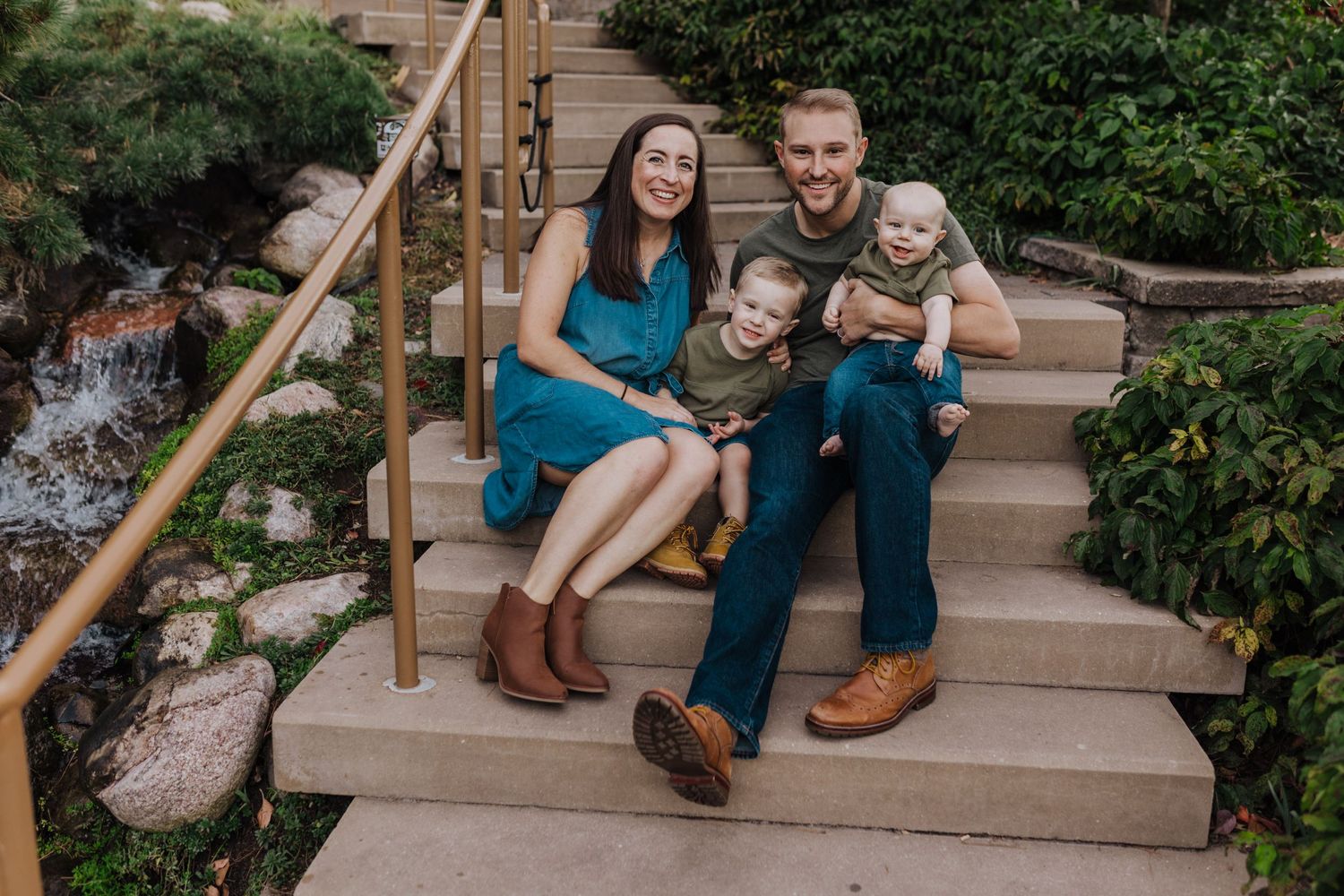 A happy family of four sits together on outdoor stone steps surrounded by garden landscaping.