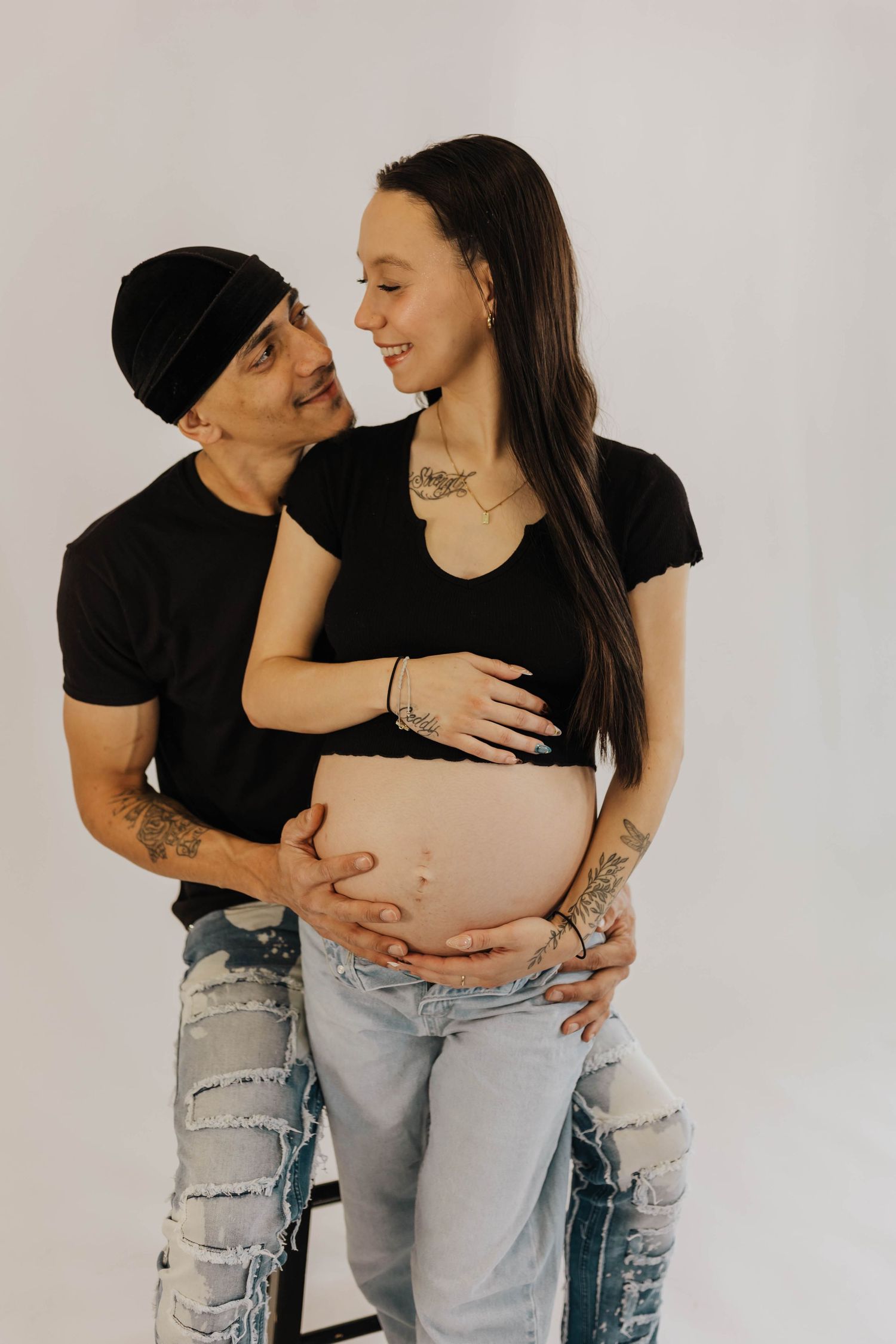 A couple wearing black shirts and ripped jeans embraces during maternity photoshoot against neutral background.