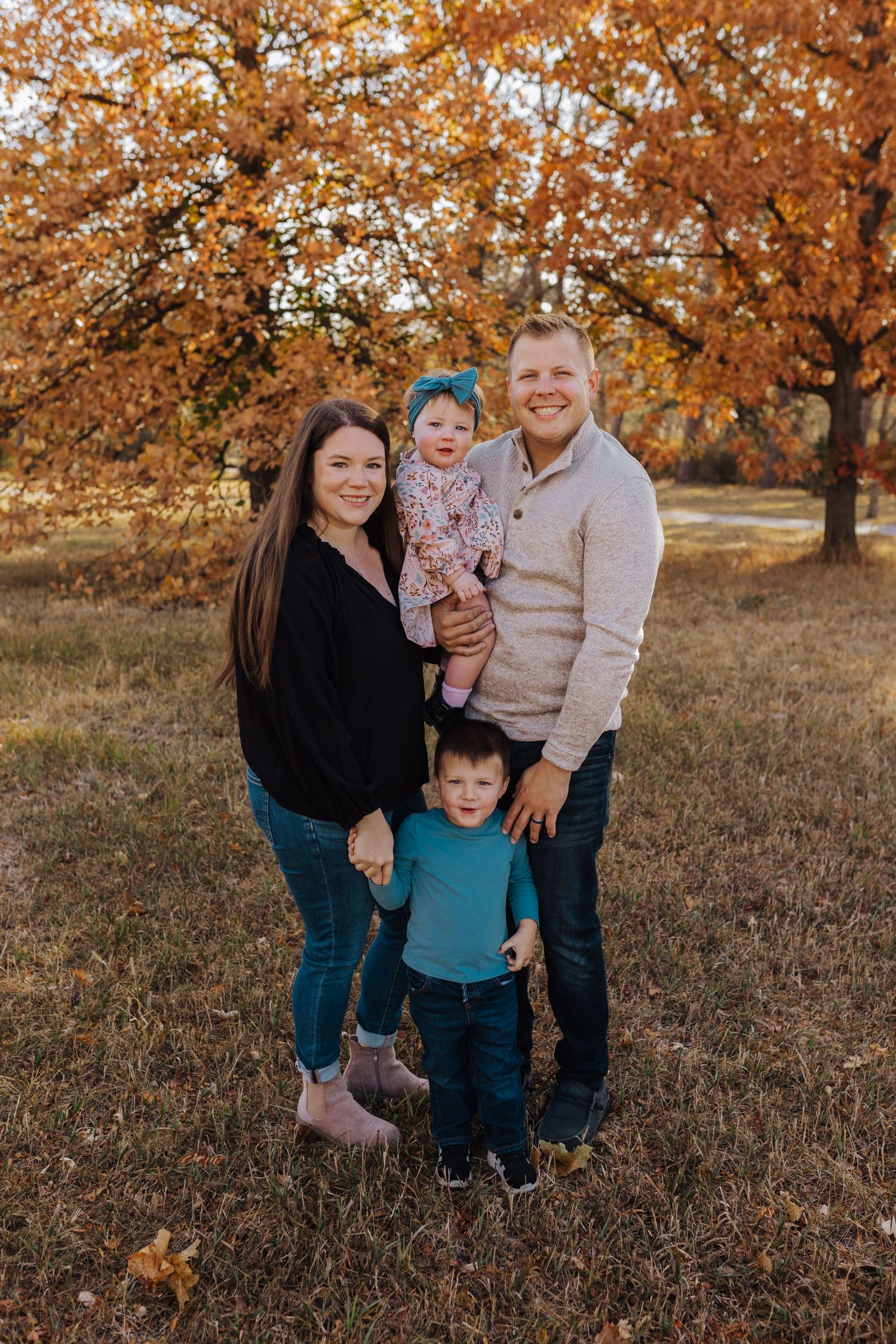 A family stands together outdoors during autumn with golden orange leaves on trees in the background.