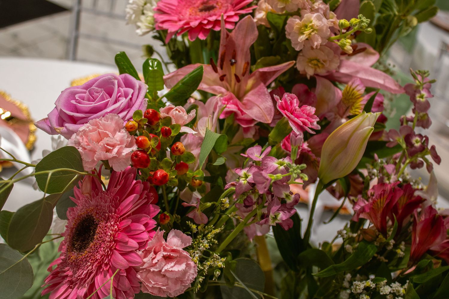 Vibrant pink floral bouquet featuring gerbera daisies, roses, lilies, and red berries with lush green foliage.