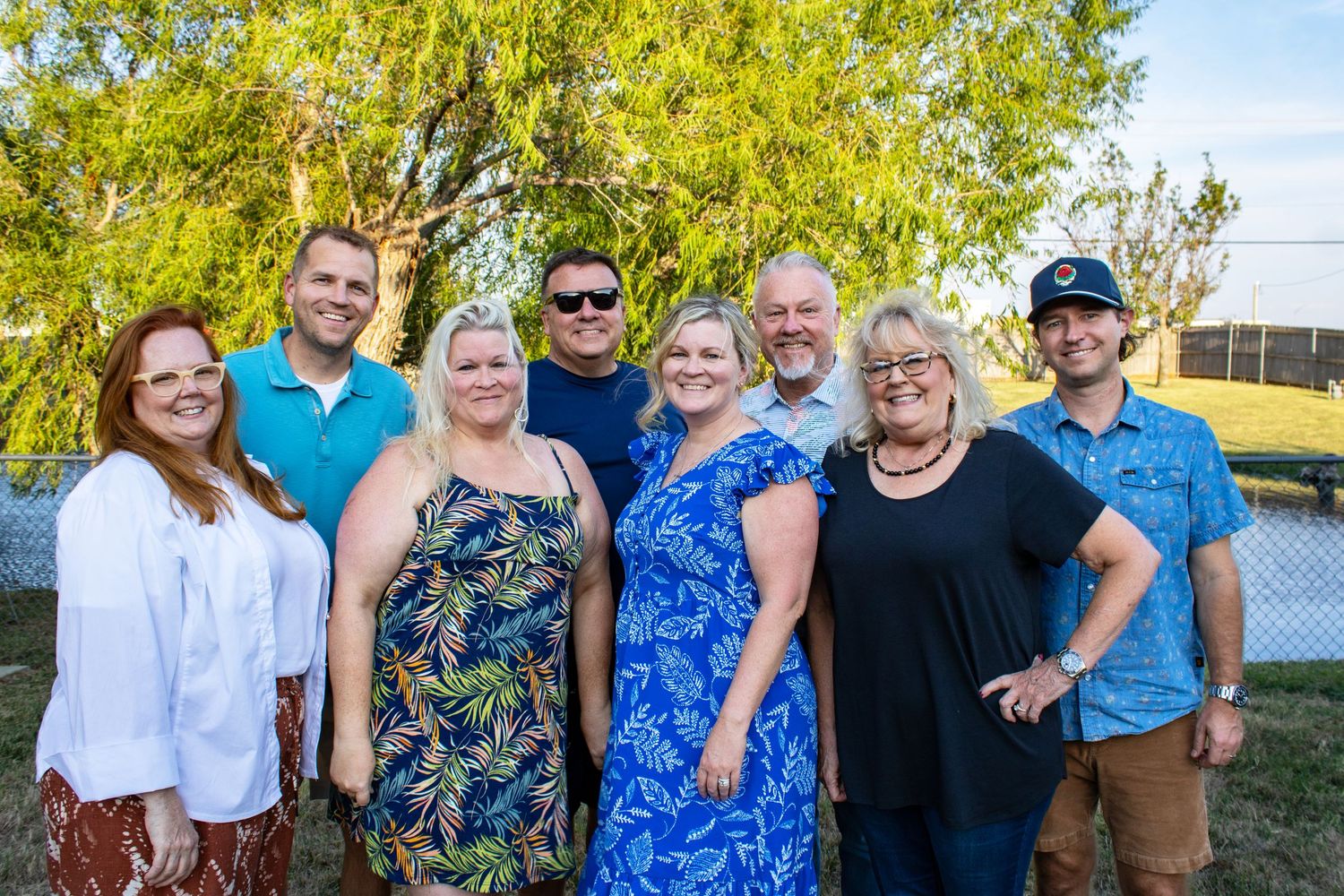 Group of eight smiling adults posing together outdoors at a casual gathering during golden hour.