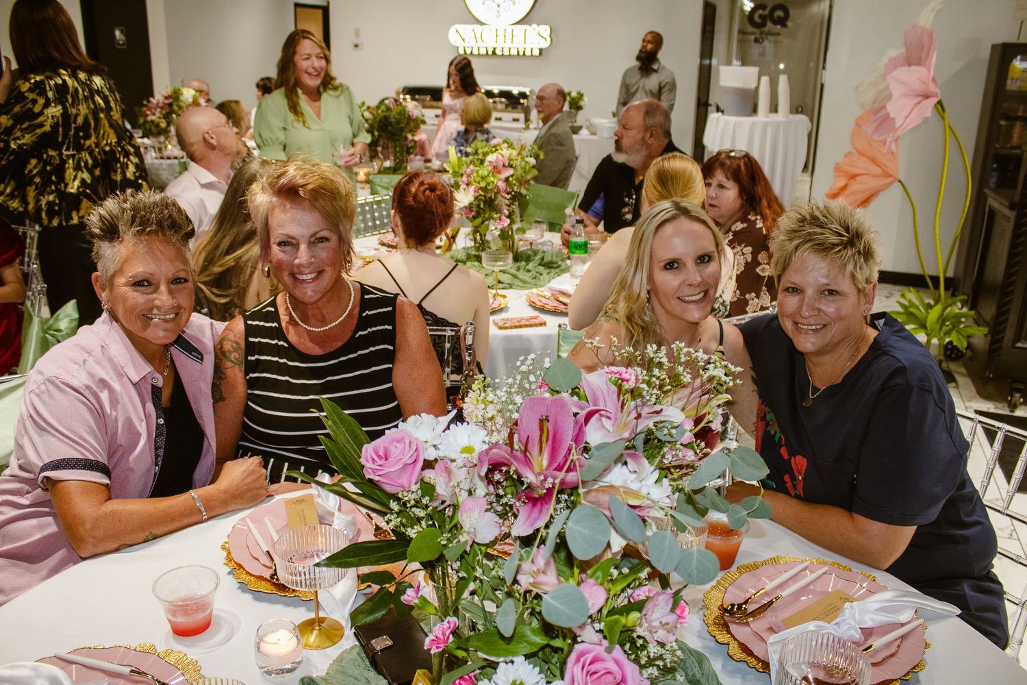 Four women smile at a floral arrangement table during an elegant flower workshop event with guests in background.