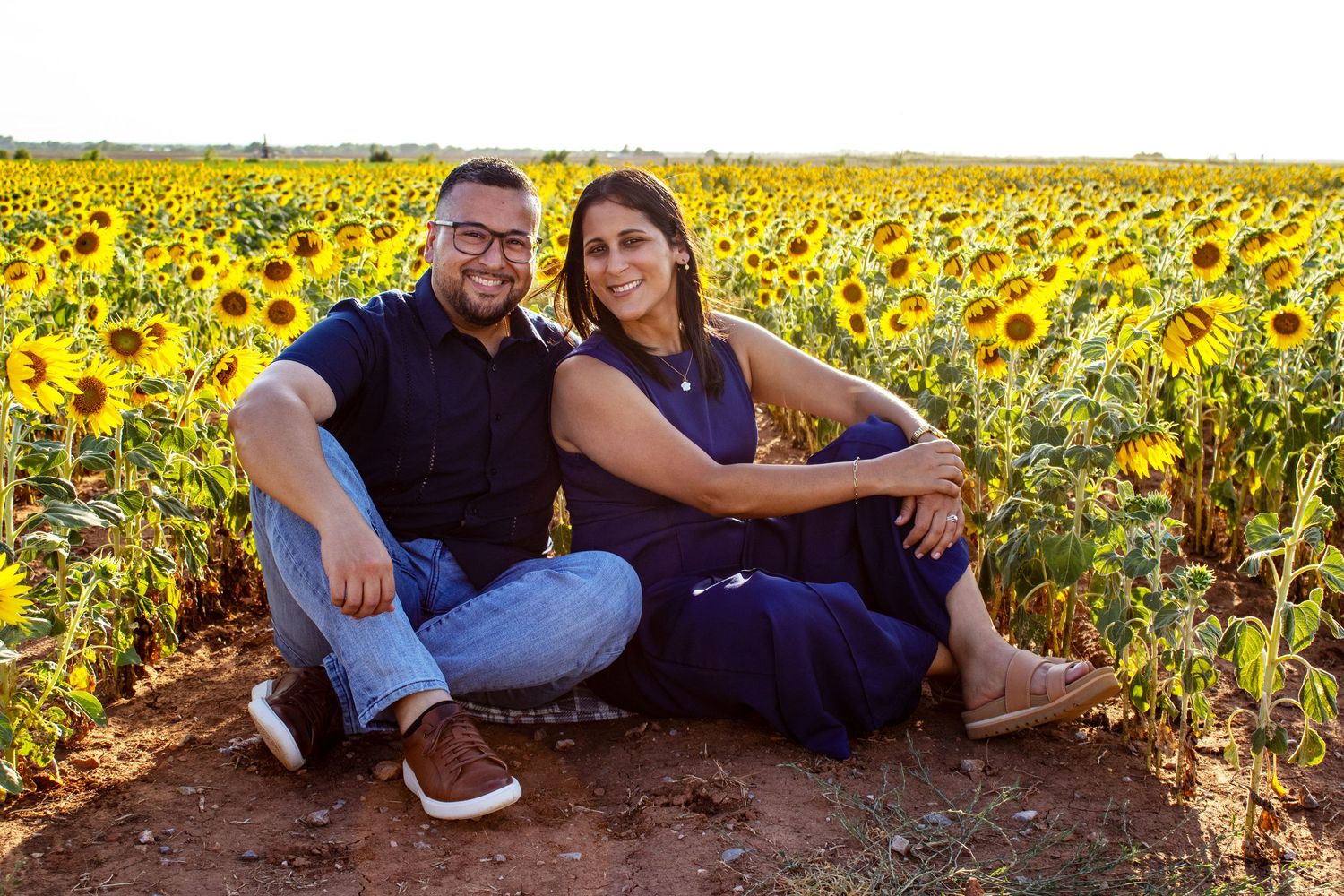 Couple smiling together in a vibrant sunflower field, wearing navy outfits, sitting on red soil during golden hour.