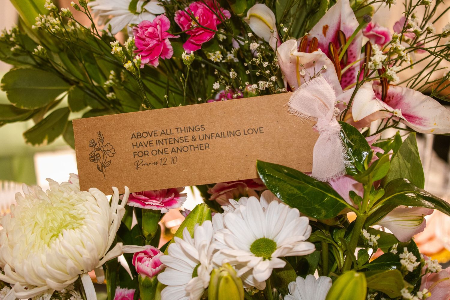 A kraft paper tag with a Bible verse about love nestled among pink and white flowers including daisies and lilies.