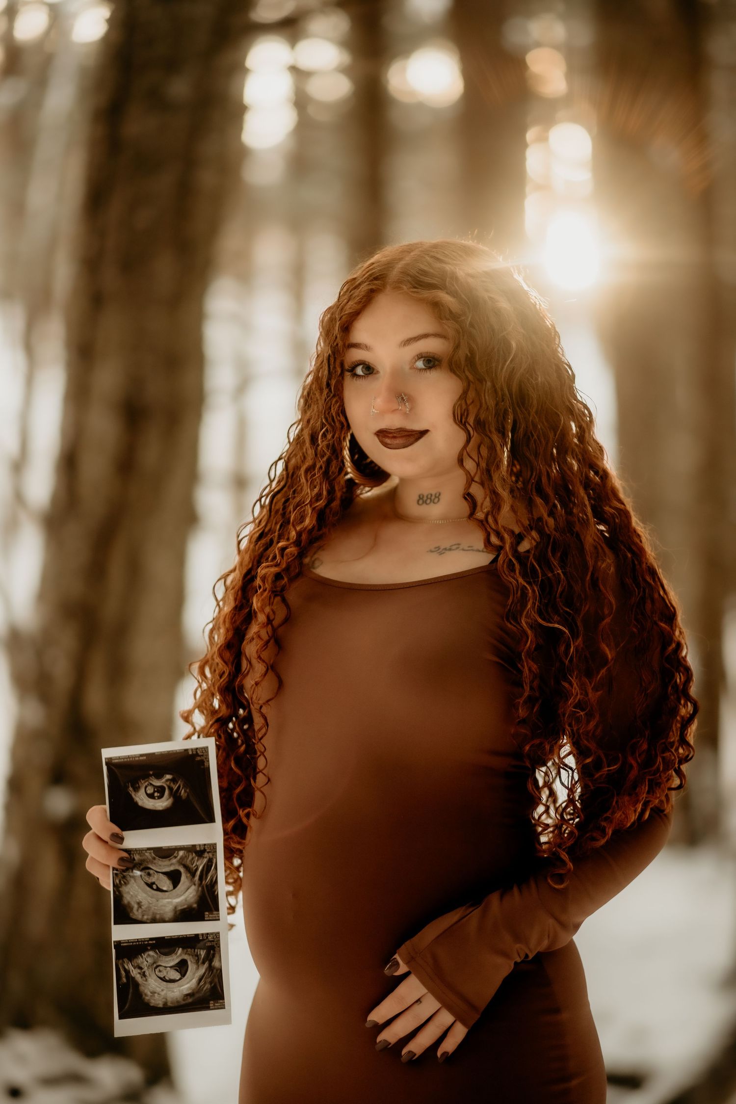 A person in a brown dress holds an ultrasound photo in a sunlit forest setting.