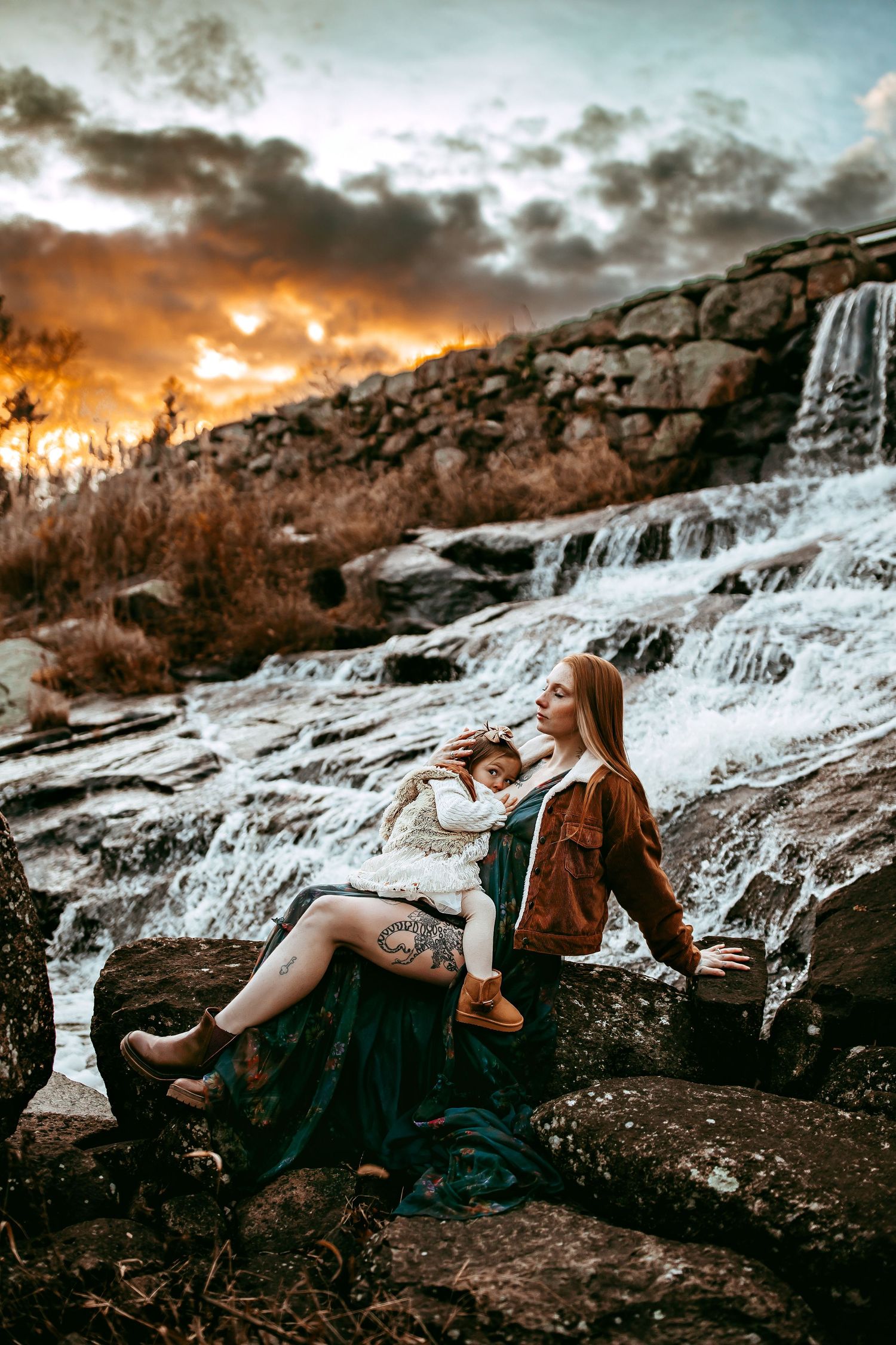 A person sits on rocky ledge drinking from bottle at scenic waterfall during dramatic orange sunset.
