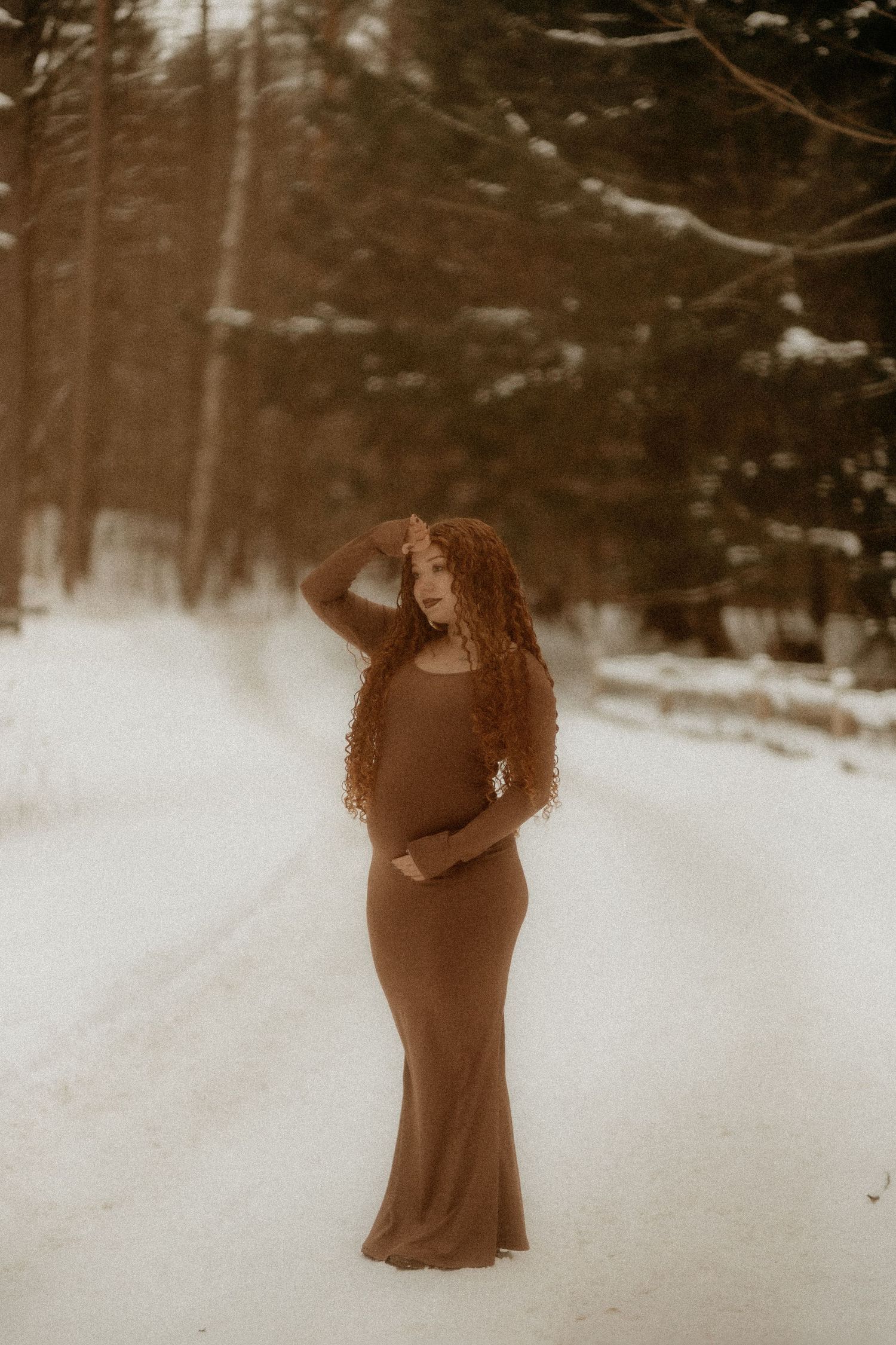 A figure in a flowing brown dress stands in a snowy winter forest setting with a dreamy, sepia-toned aesthetic.