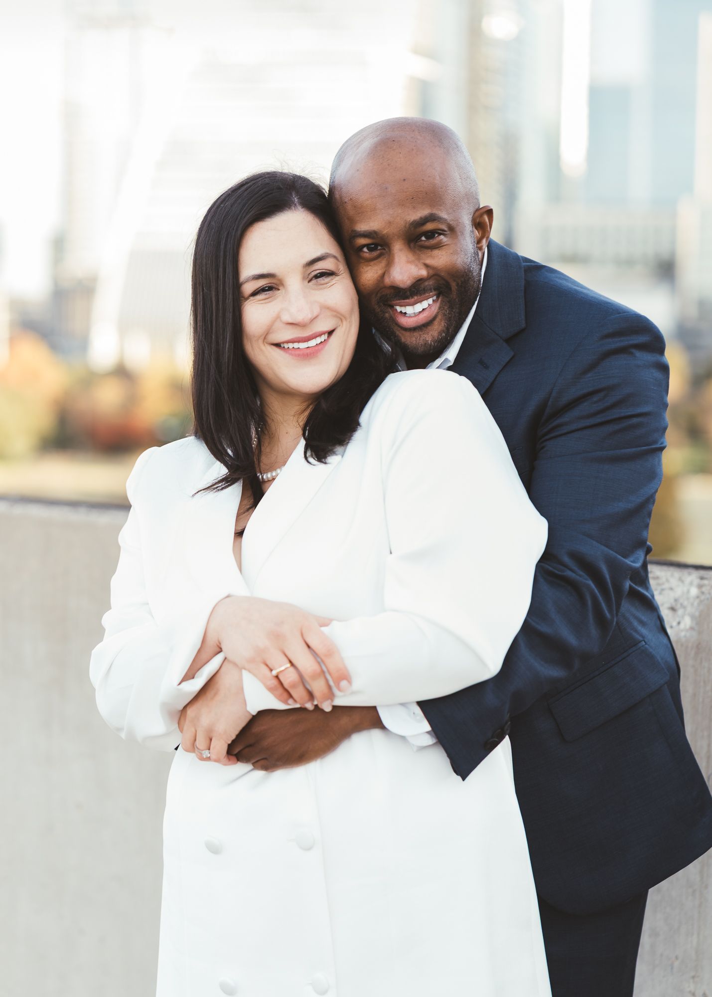Couple smiling toward the camera on a rooftop, with the man embracing his partner from behind.