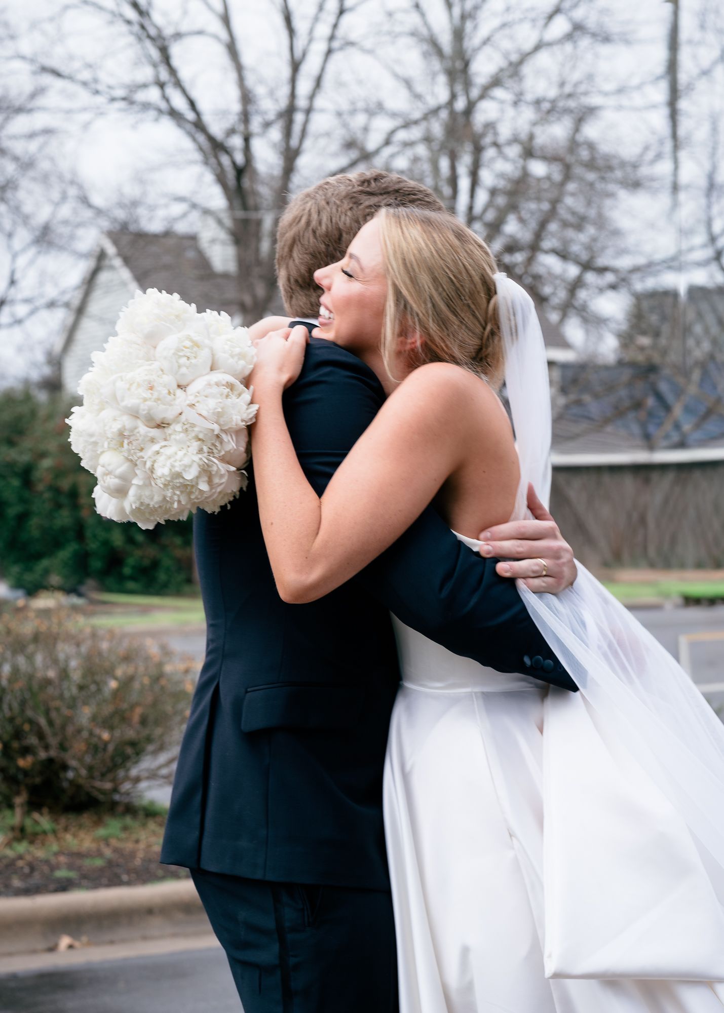 Bride smiling as she hugs her groom outdoors, holding a white bouquet over his shoulder.