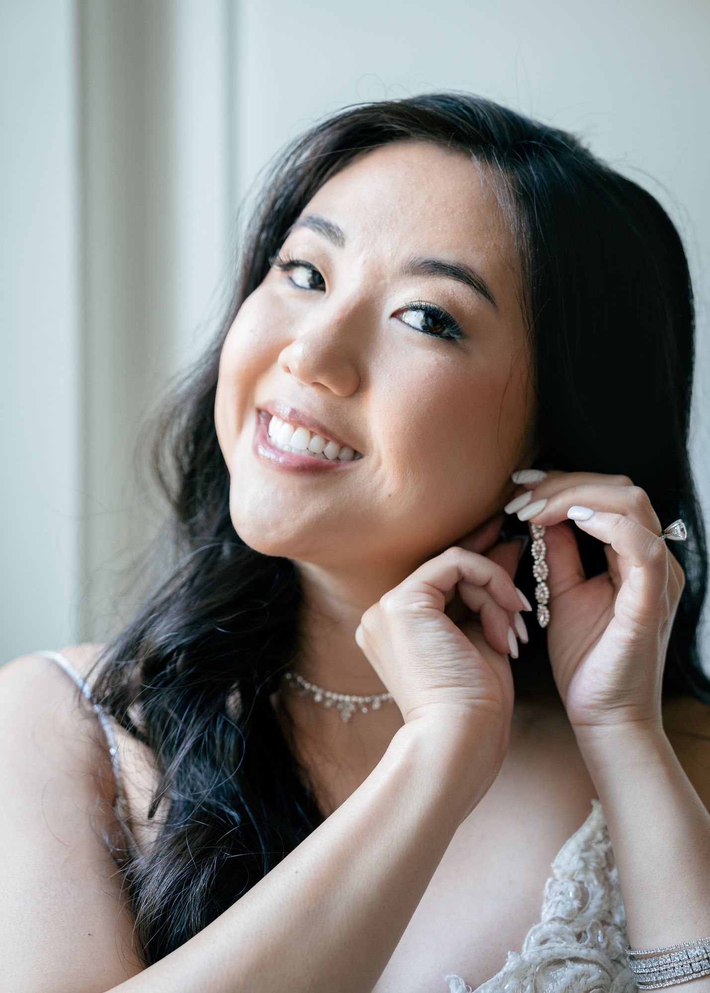 Smiling bride adjusting a dangling earring beside a window, wearing a lace wedding dress.