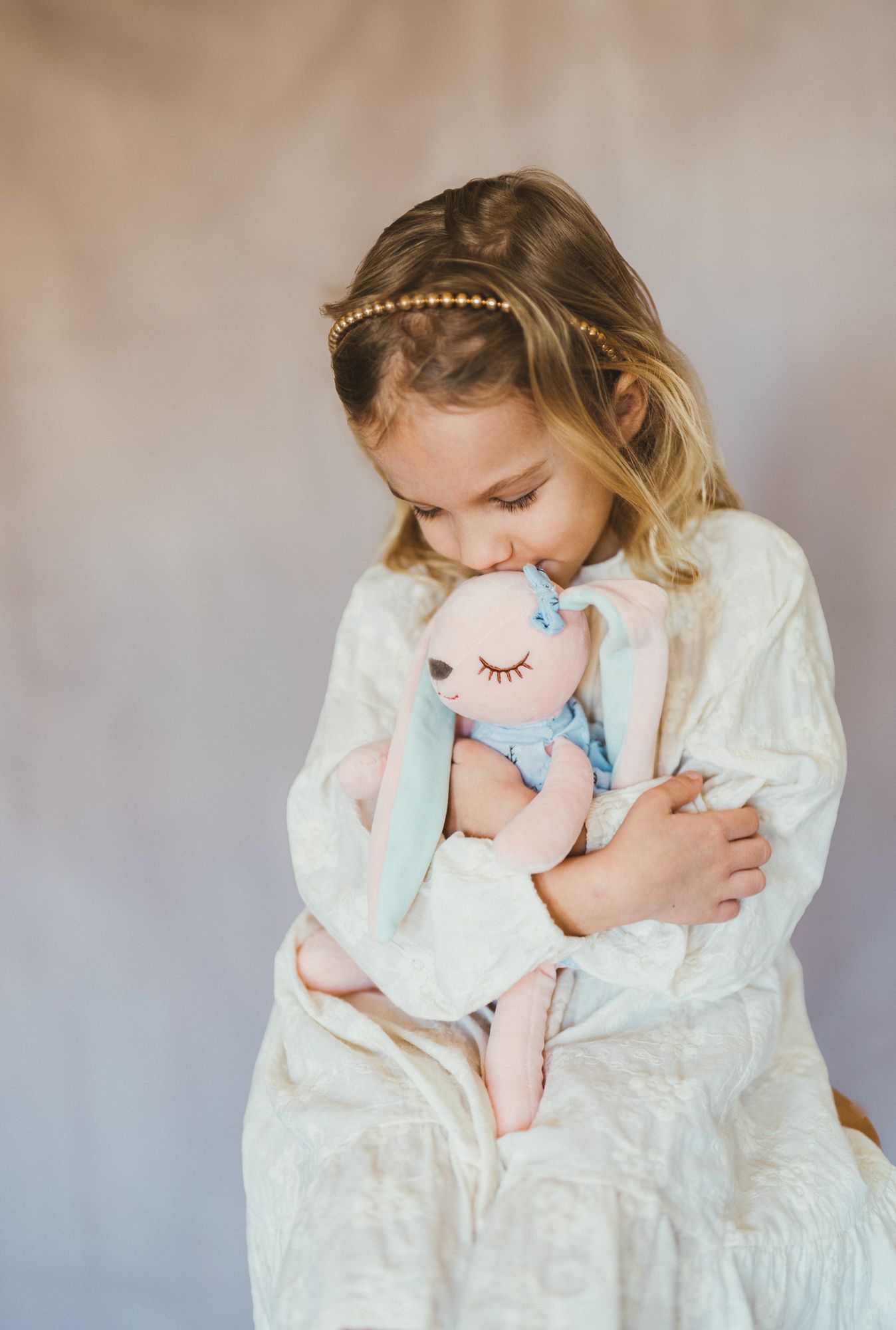 Girl in a cream dress looking down as she hugs a pink stuffed rabbit.