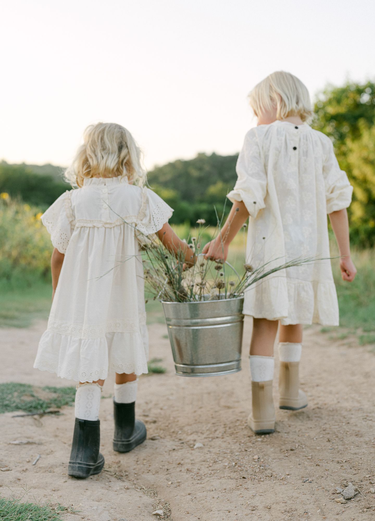 Two girls in white dresses and rain boots walking away together, sharing a metal bucket filled with grasses.