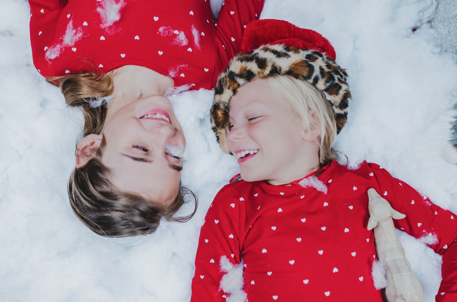 Two girls in matching red holiday pajamas laughing together while lying in faux snow during a winter photography session in Austin, Texas.