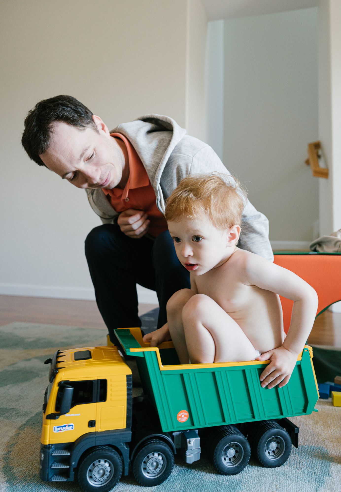 Father leaning toward his toddler seated inside a yellow and green toy dump truck at home.