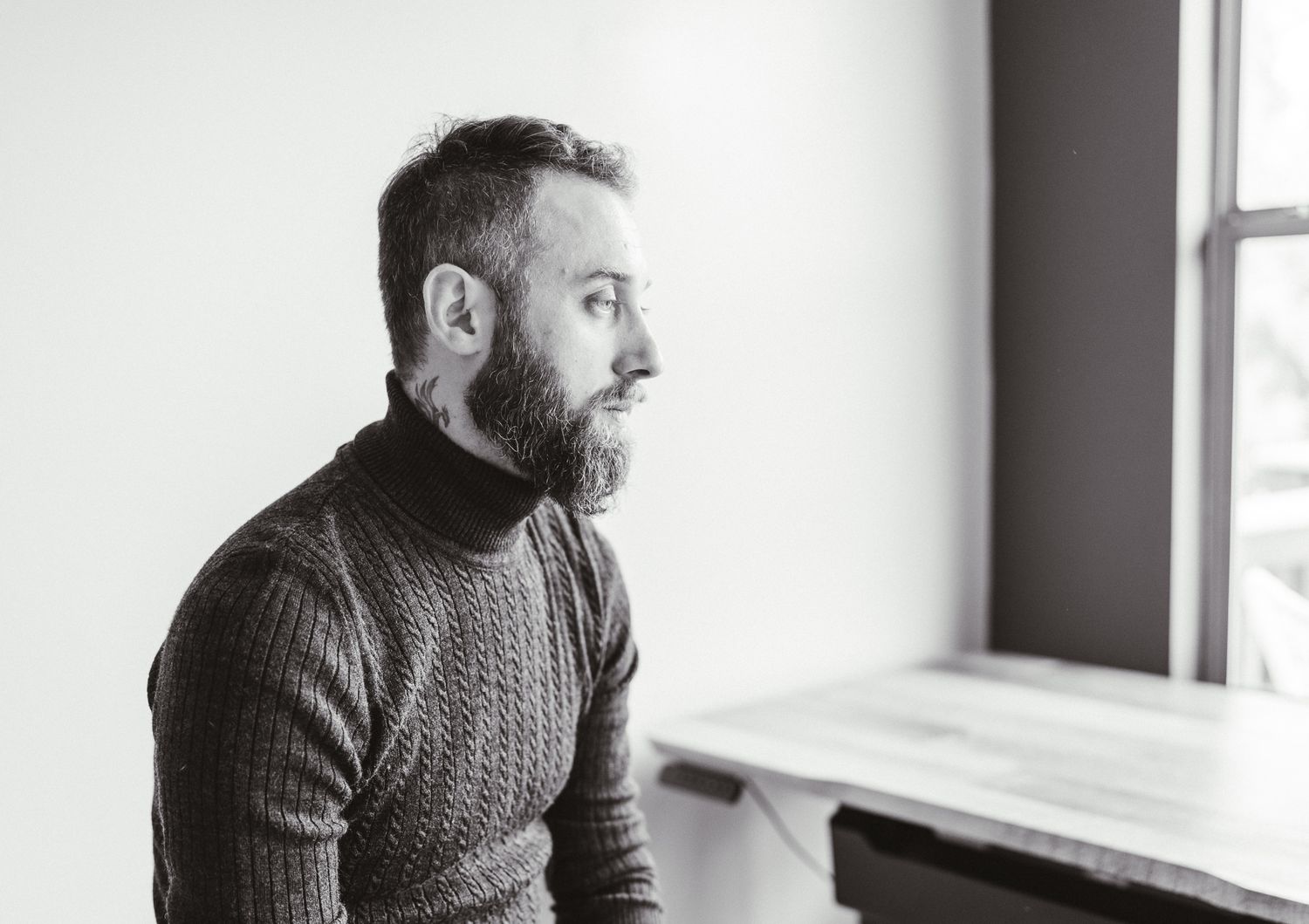 Black-and-white portrait of a bearded man in a knit sweater gazing toward a window.