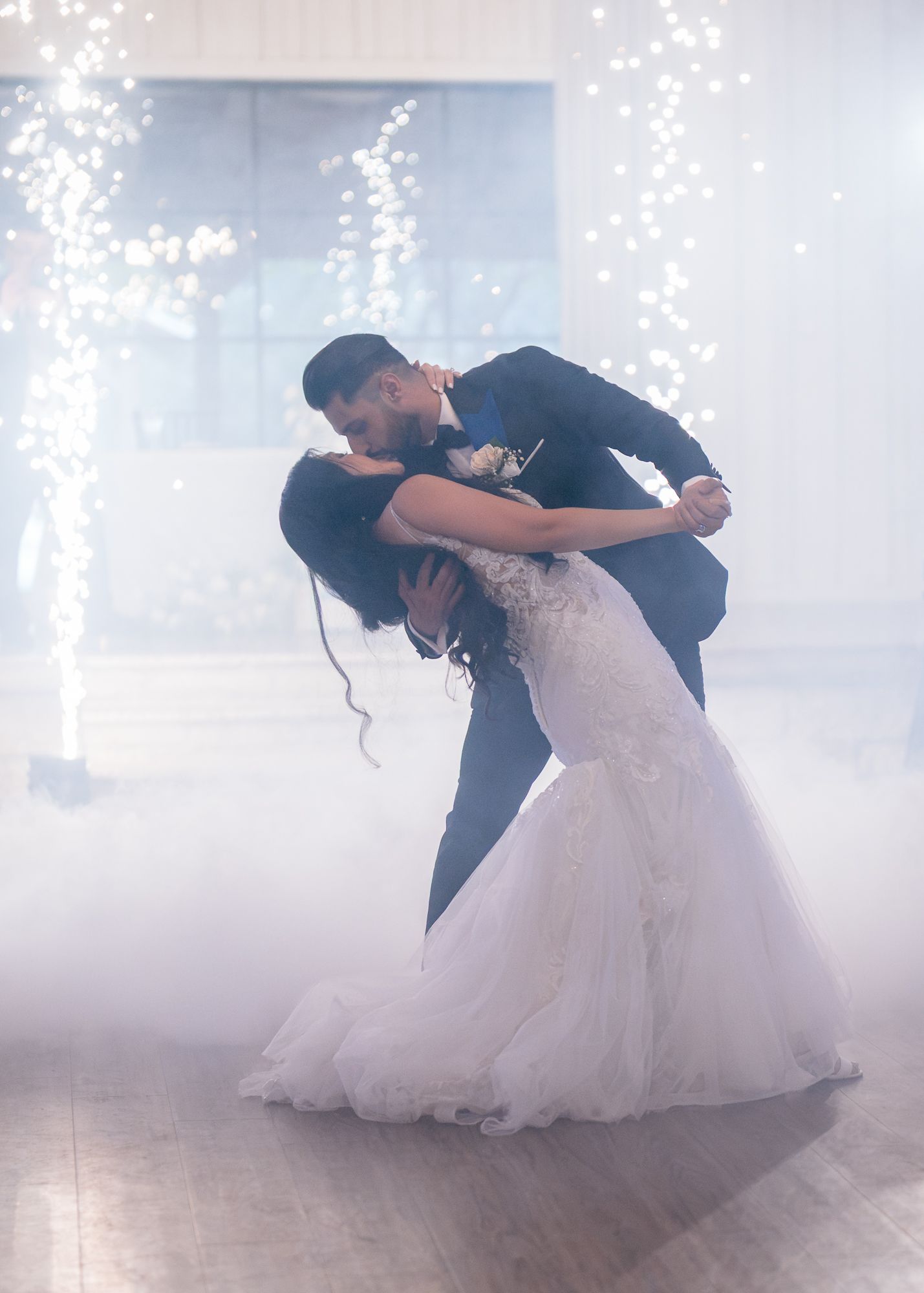 Groom dipping and kissing his bride amid rising sparks and low mist on the dance floor.