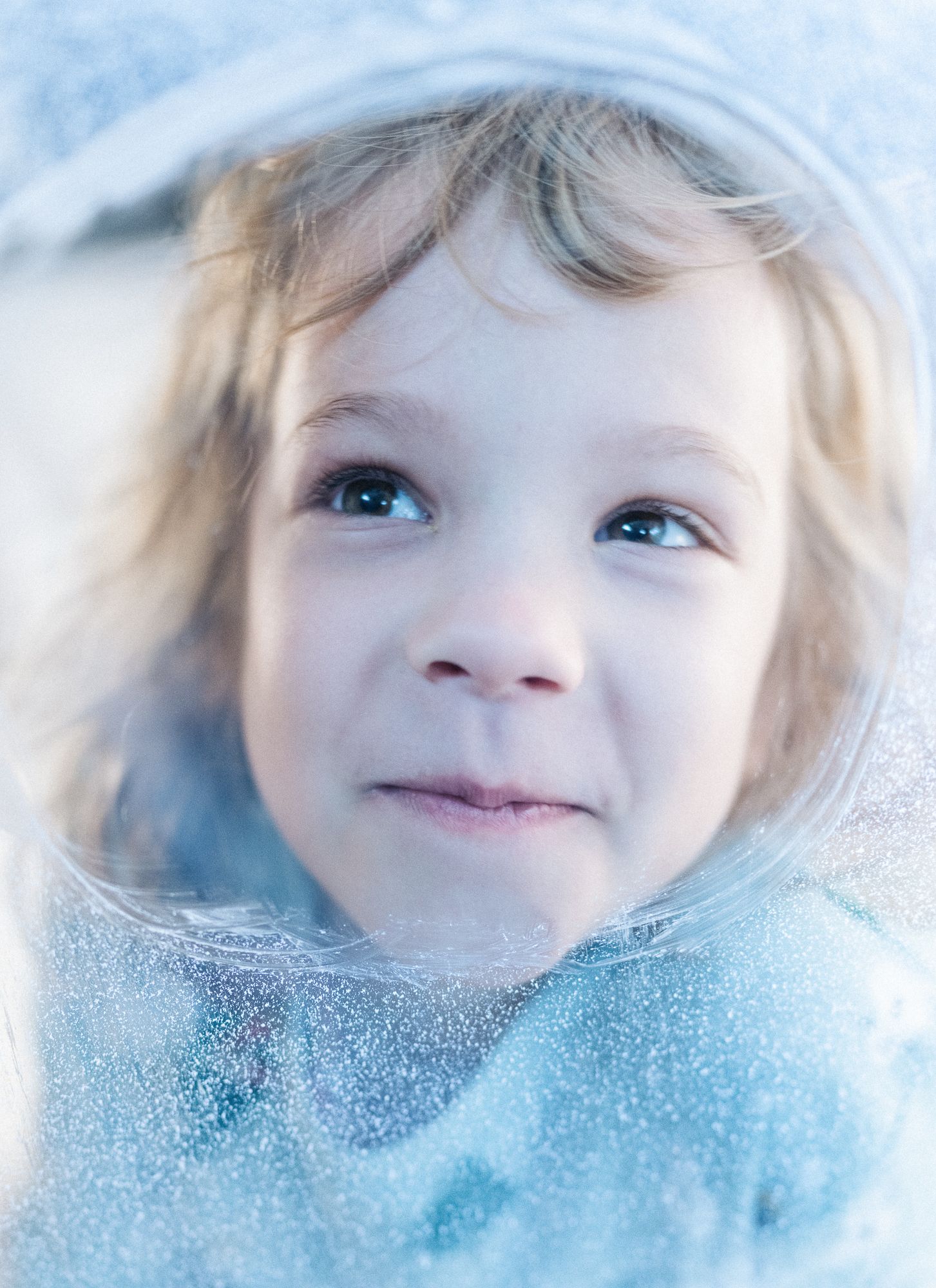 Close-up winter portrait of a young girl framed by frosted ice in soft blue light.