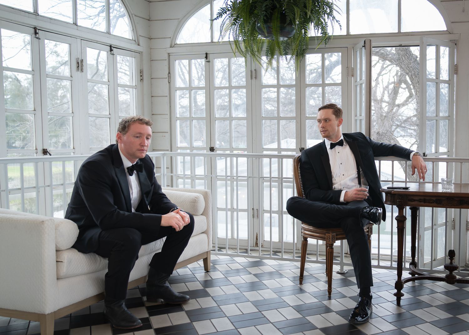 Two men in tuxedos sharing a quiet moment in a bright glass-enclosed room