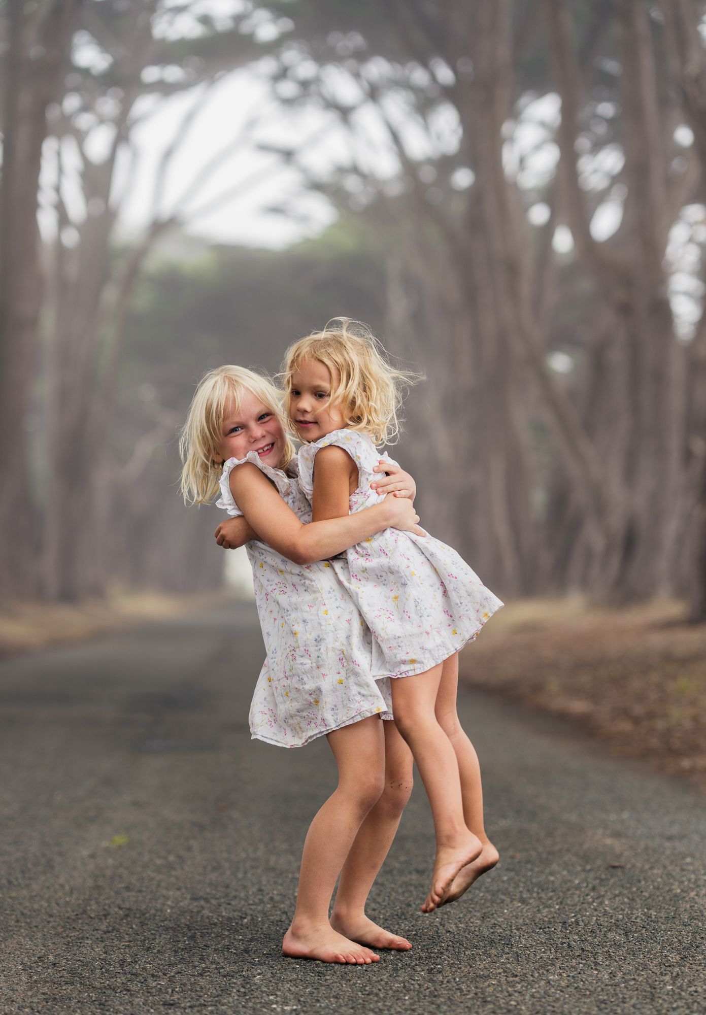 Barefoot sisters embracing on a tree-lined path, the older sister lifting the younger.