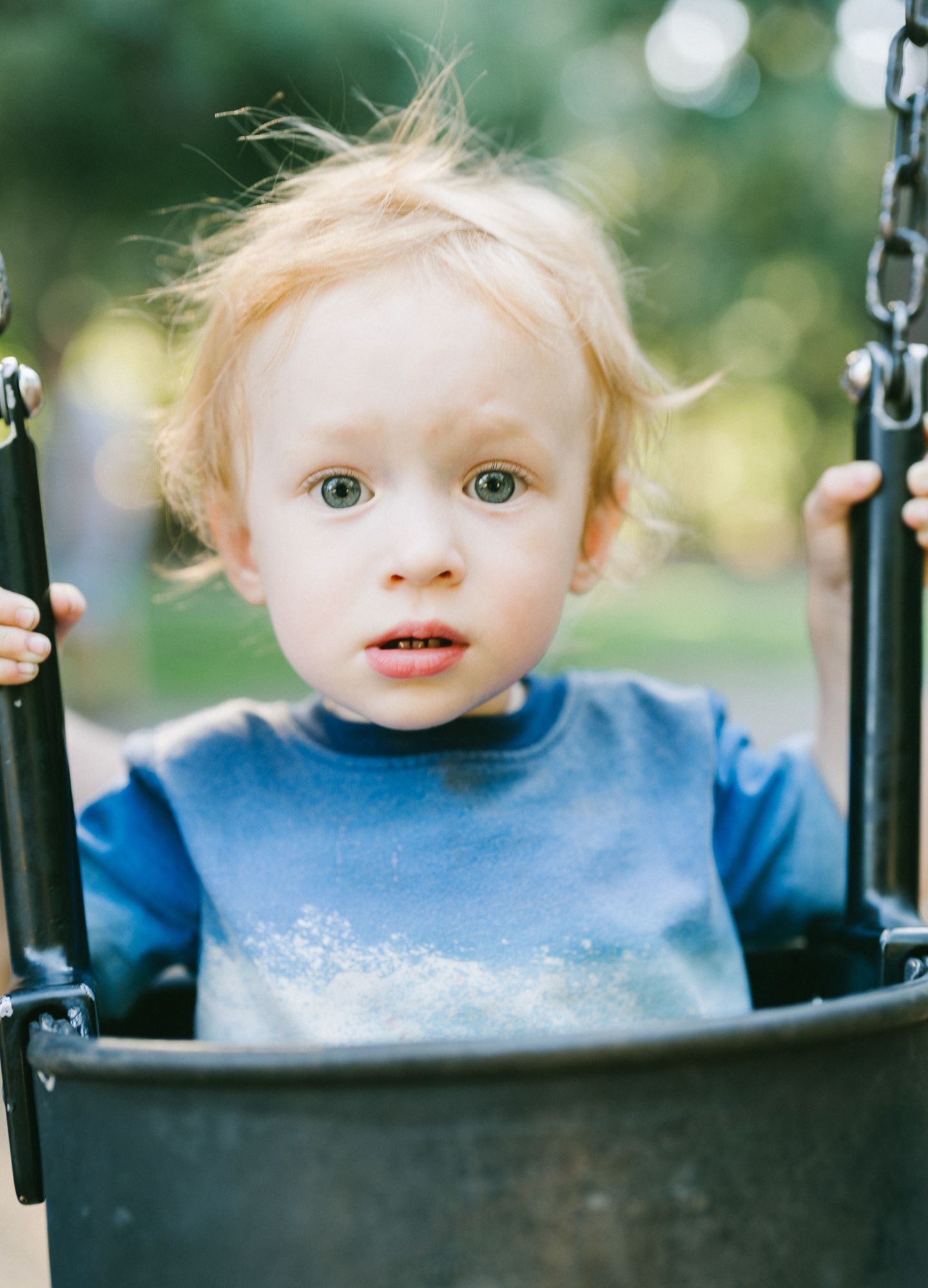 Toddler in a blue shirt holding the sides of a playground swing and looking at the camera.