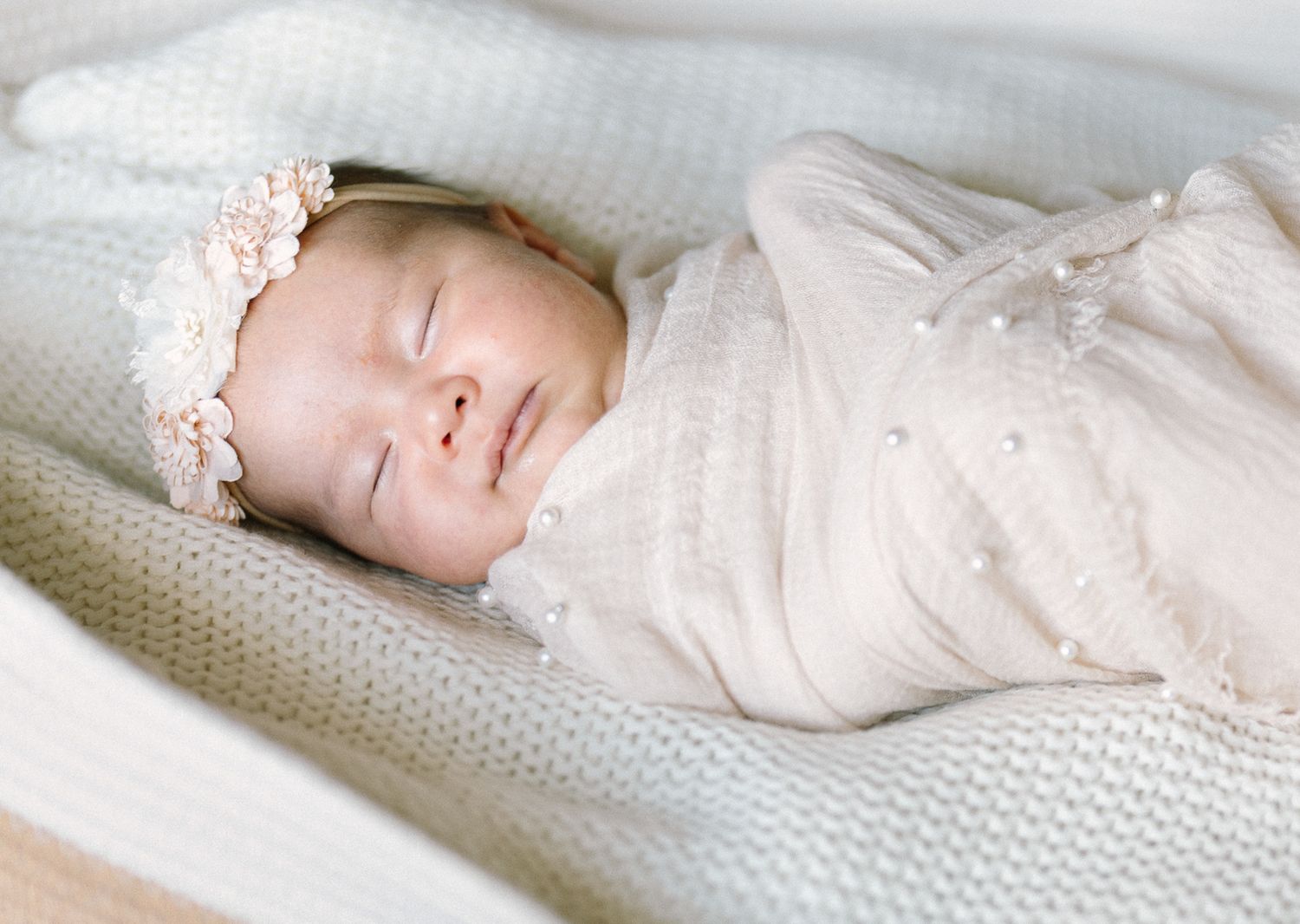 Sleeping newborn wearing a floral headband, swaddled in a cream wrap on a knitted blanket.