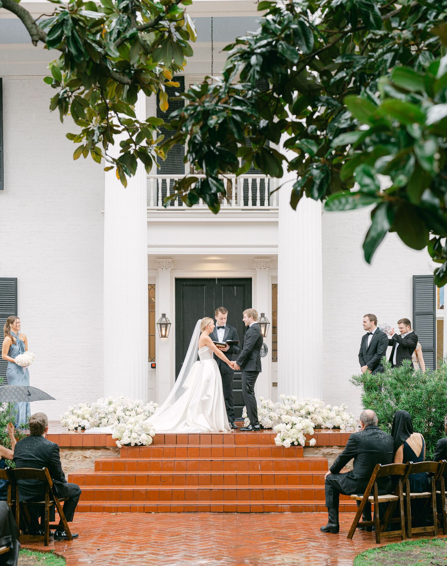 Couple exchanging vows on the brick steps of a white-columned mansion, with guests seated nearby.