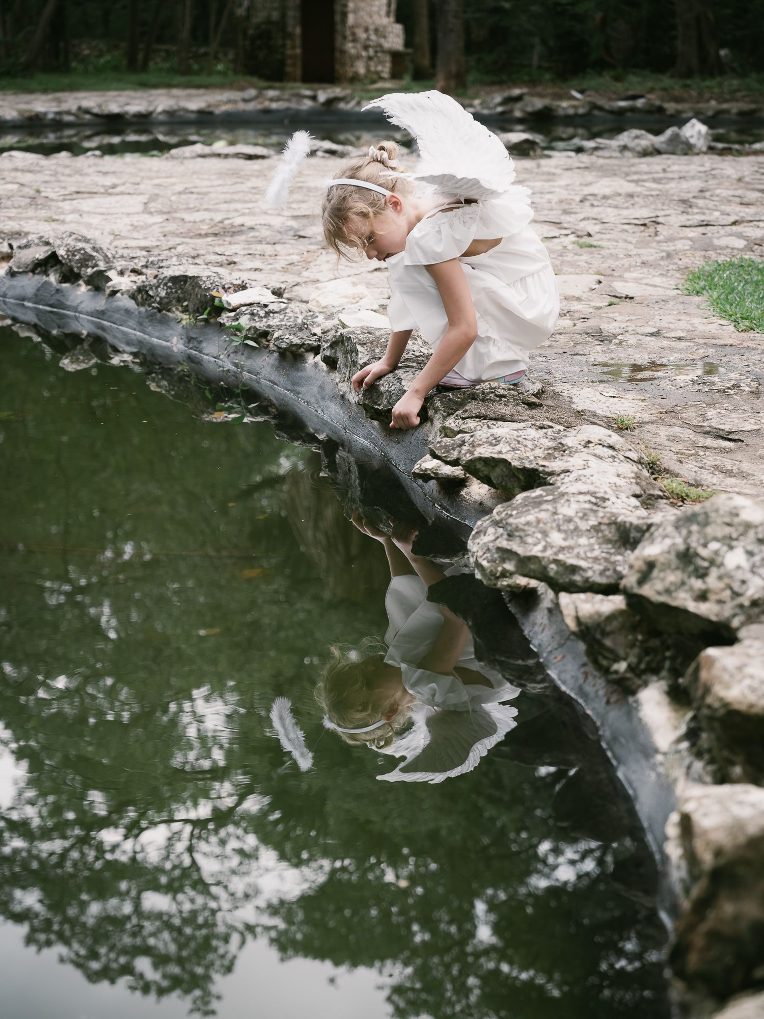 Girl wearing white angel wings crouching beside a pond, with her reflection visible in the water.