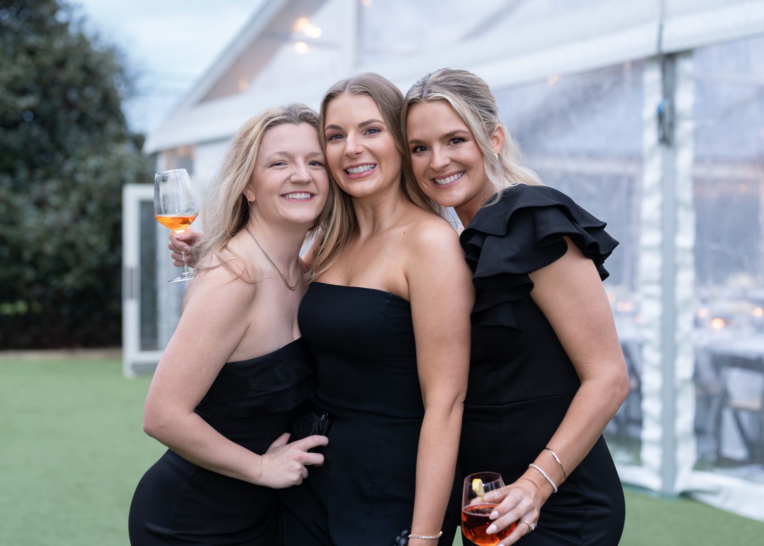 Three women in black dresses smiling together with drinks at an outdoor celebration