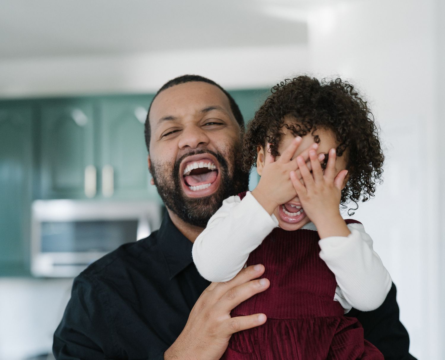 Father laughing while holding his child, who covers their eyes with both hands.