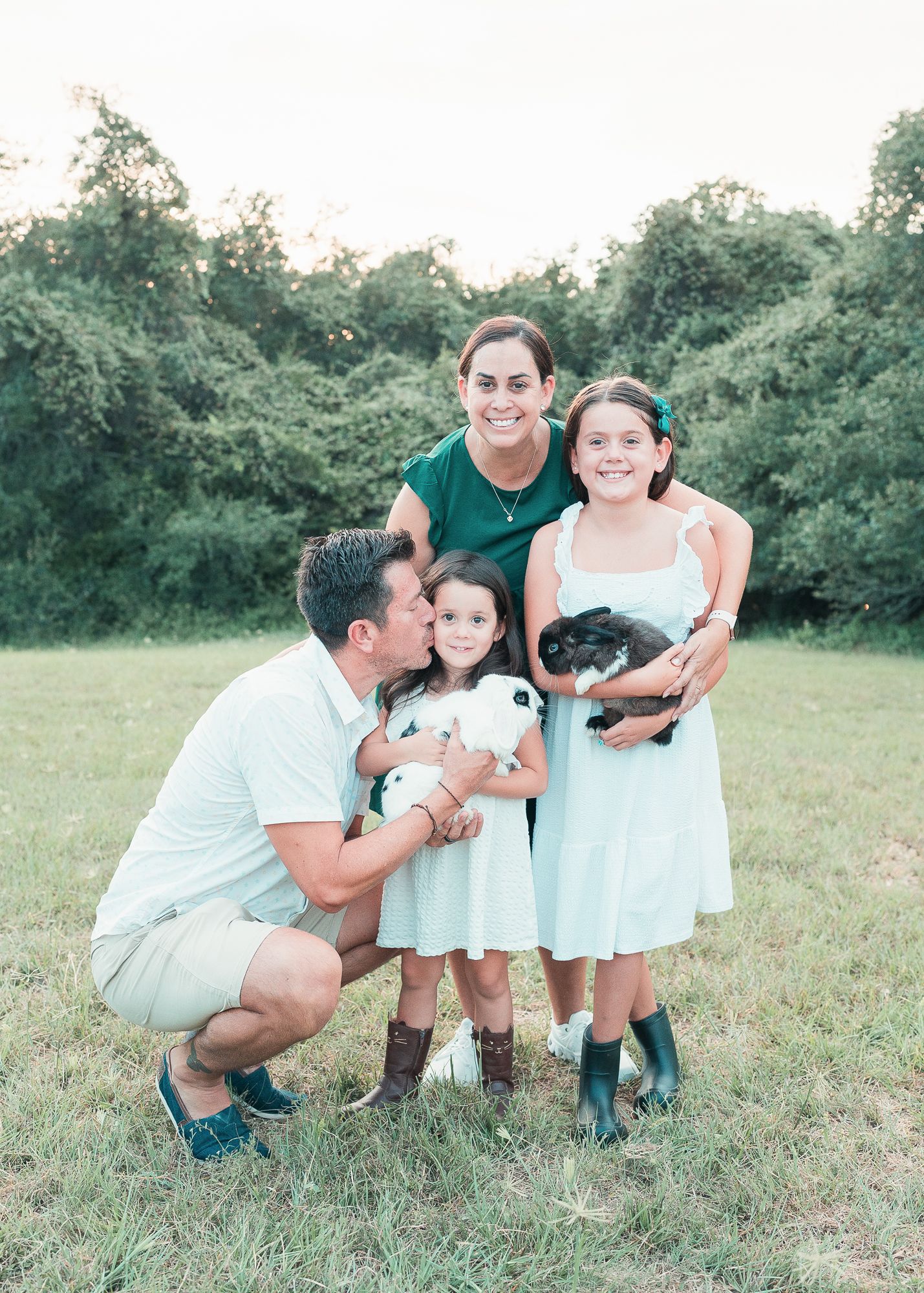 Parents and two daughters holding rabbits outdoors, with the father kissing his younger daughter’s cheek.