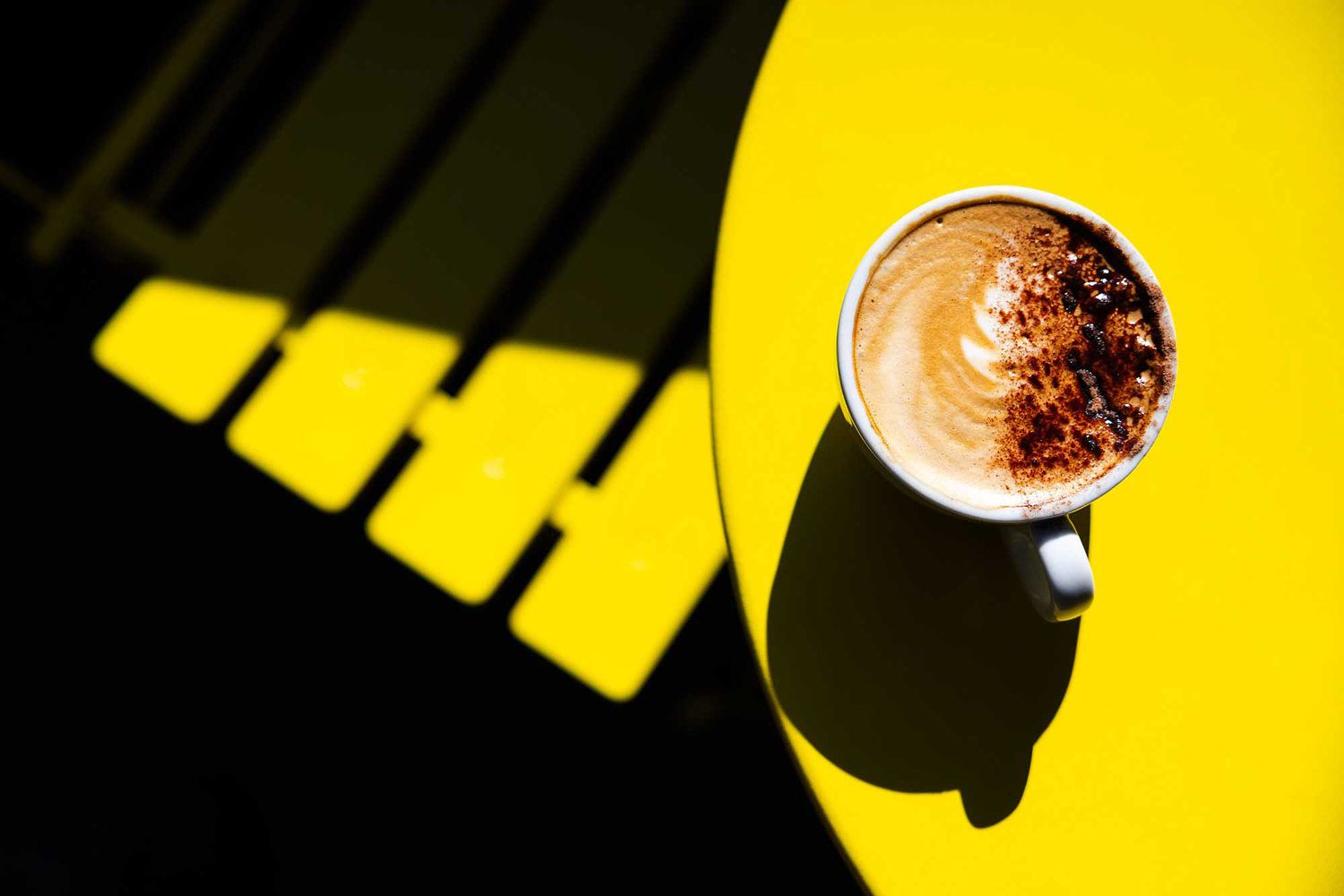 Cup of cappuccino on a bright yellow café table, captured in dramatic sunlight with bold shadows and minimalist composition.