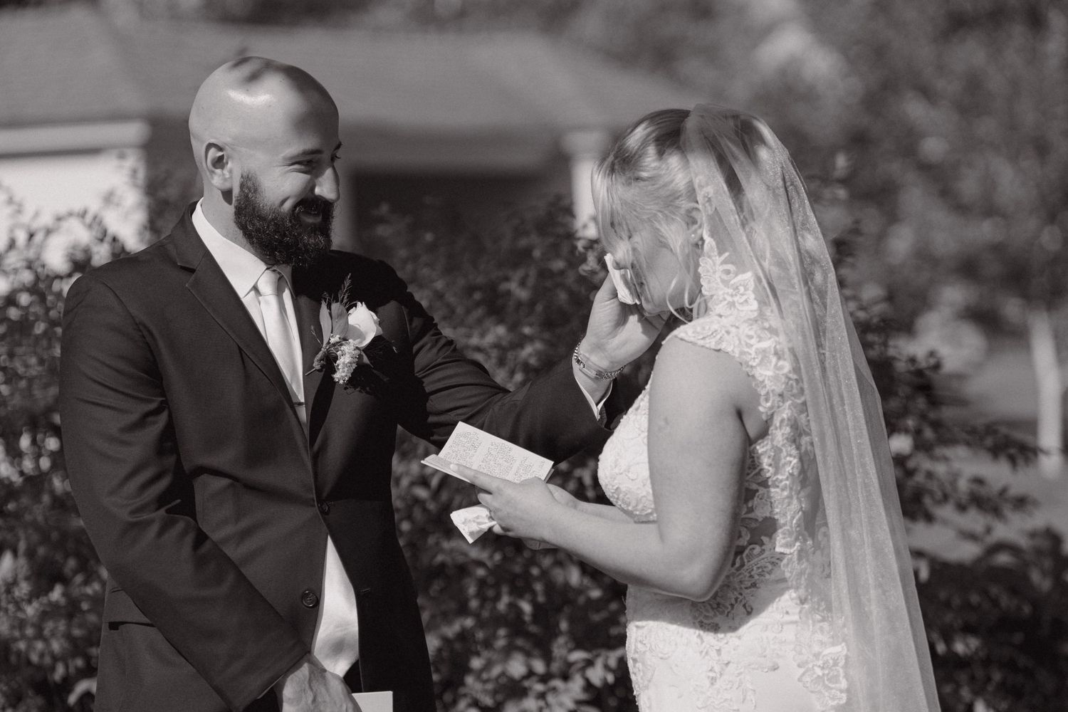 Black and white photo of bride reading vows while groom smiles tenderly during outdoor wedding ceremony.