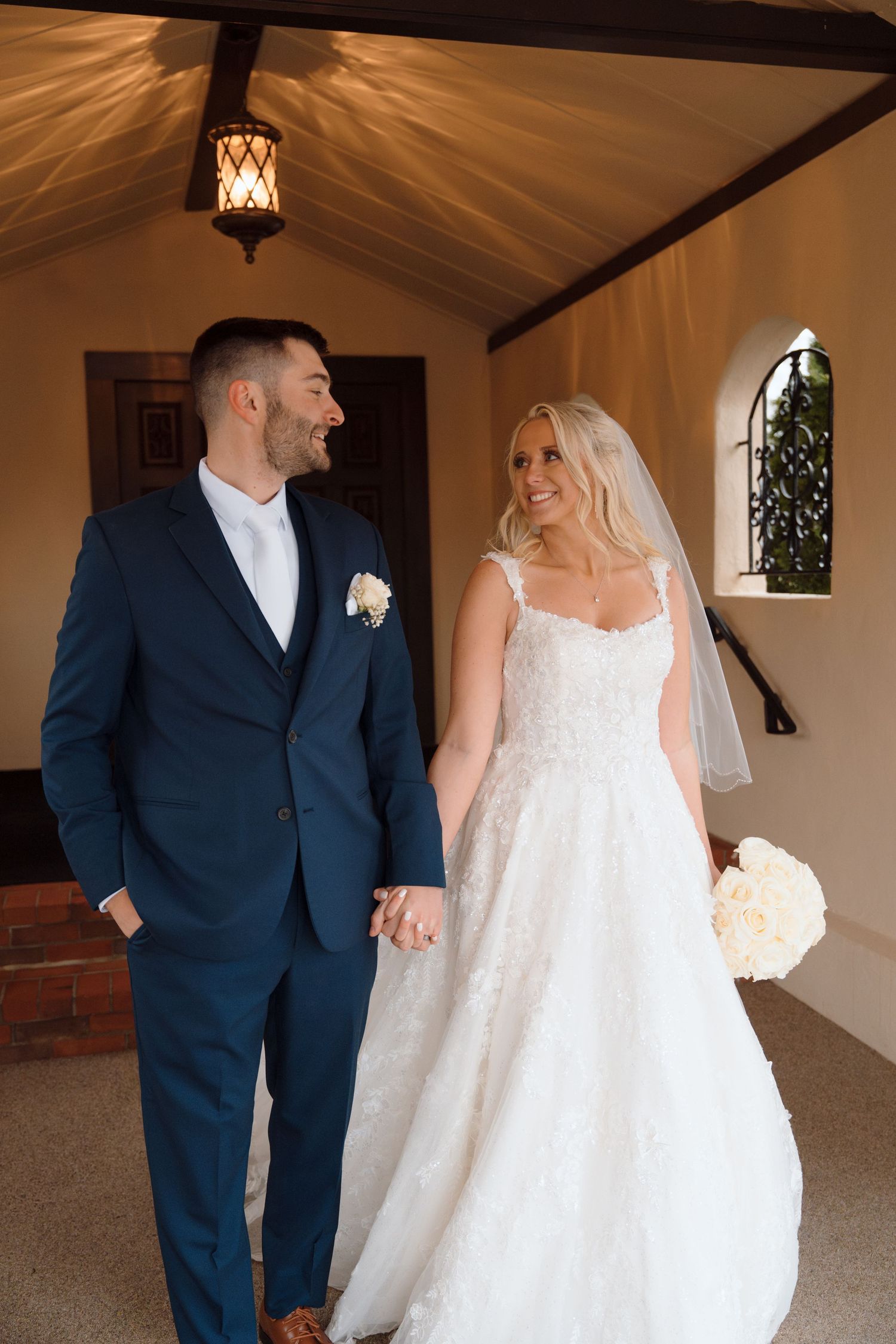 Bride in white lace gown holding bouquet walks hand in hand with groom in navy suit inside a chapel.