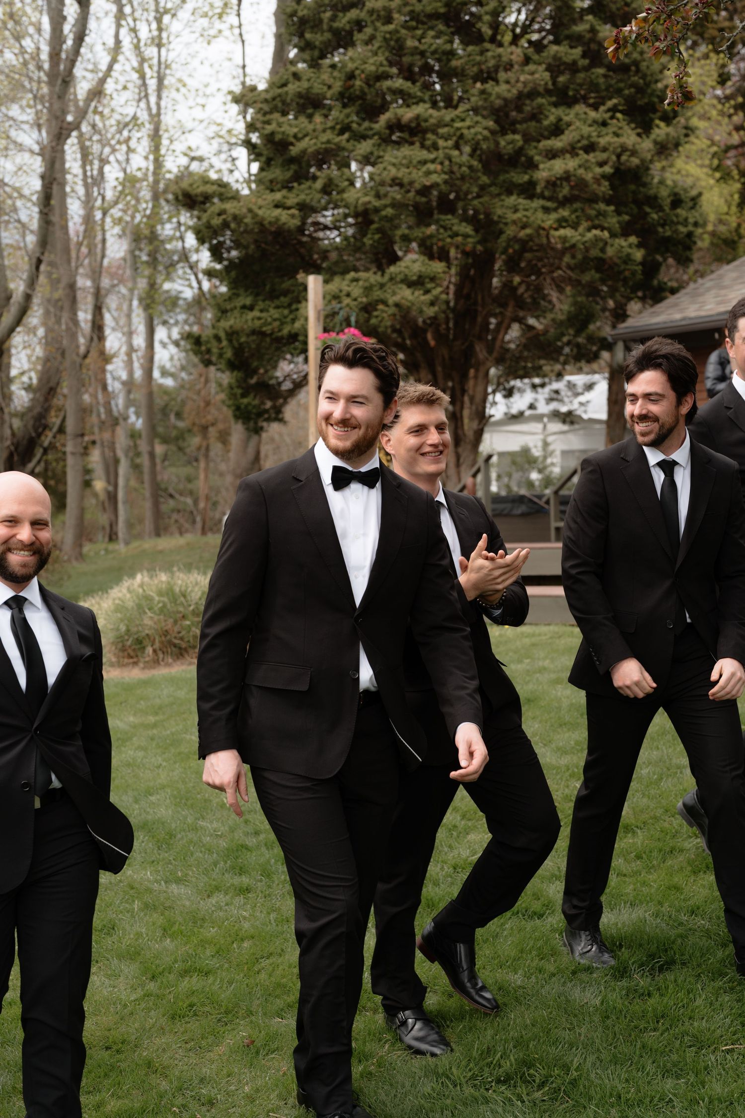 Groomsmen in black tuxedos laughing and walking outdoors on green grass with trees in background at a wedding.