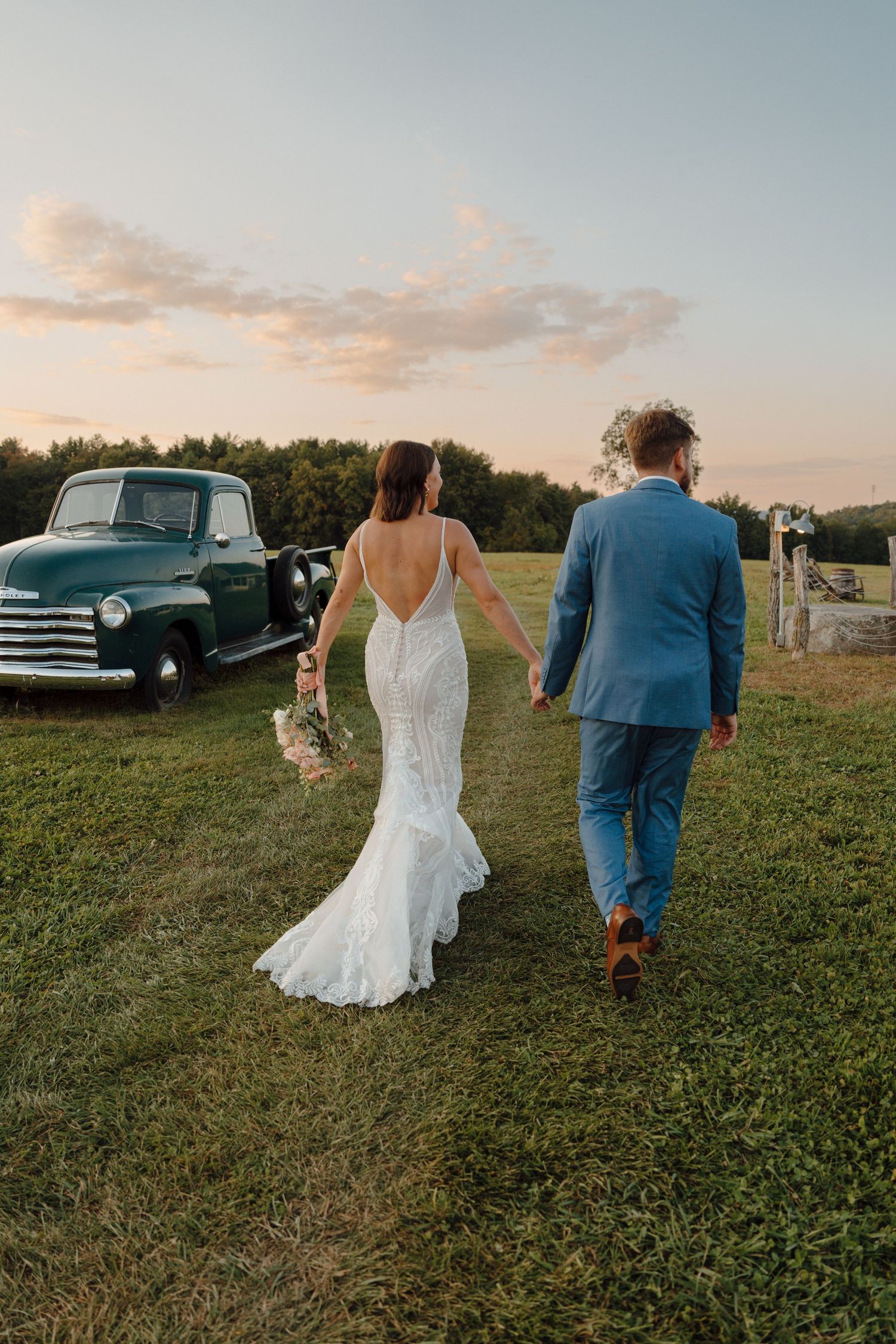 A couple walking together on a grassy hill at sunset with vintage cars parked in the background.